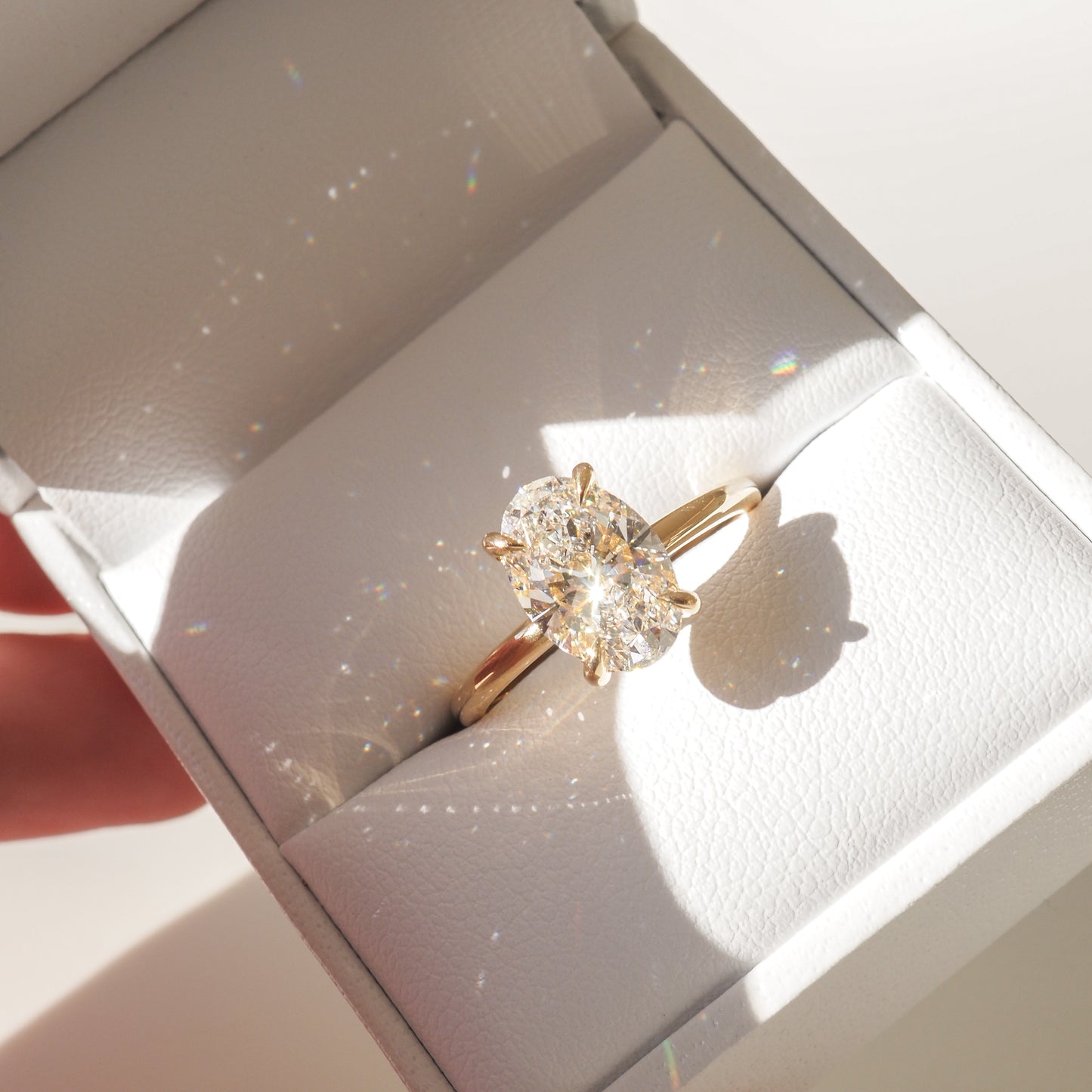 A stunning oval diamond ring in a gold setting sits in an elegant white jewellery box, illuminated by sunlight, casting sparkling reflections.