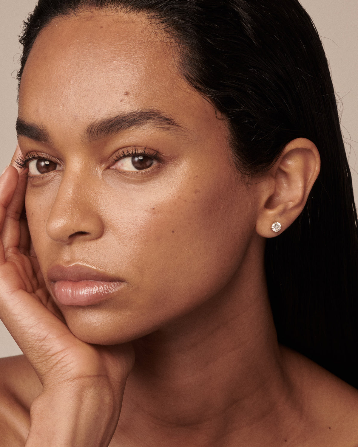 A close-up portrait of a woman with dark hair, wearing a small round silver stud earring with a clear diamond, set against a warm beige background. [text overlay: The Clara Studs in 1.5ct (0.75ct each)]