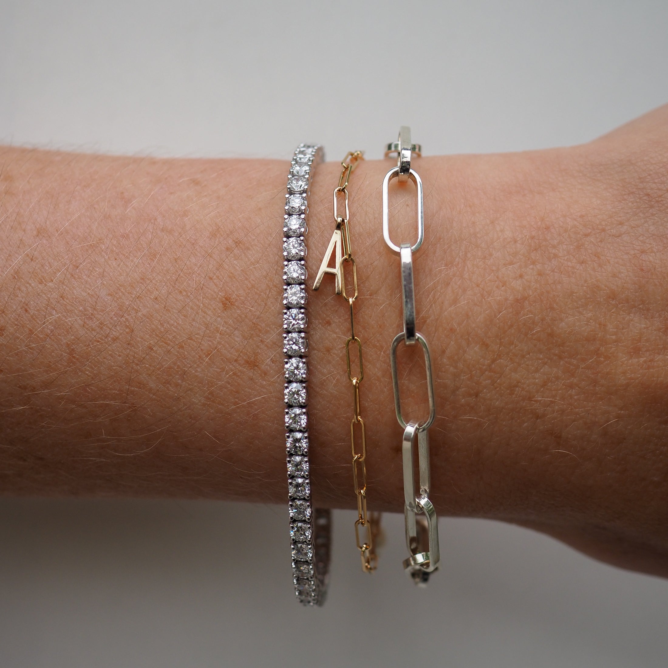 Close-up of a wrist wearing three luxury bracelets: a diamond tennis bracelet, a gold chain with charm, and a silver link chain bracelet.