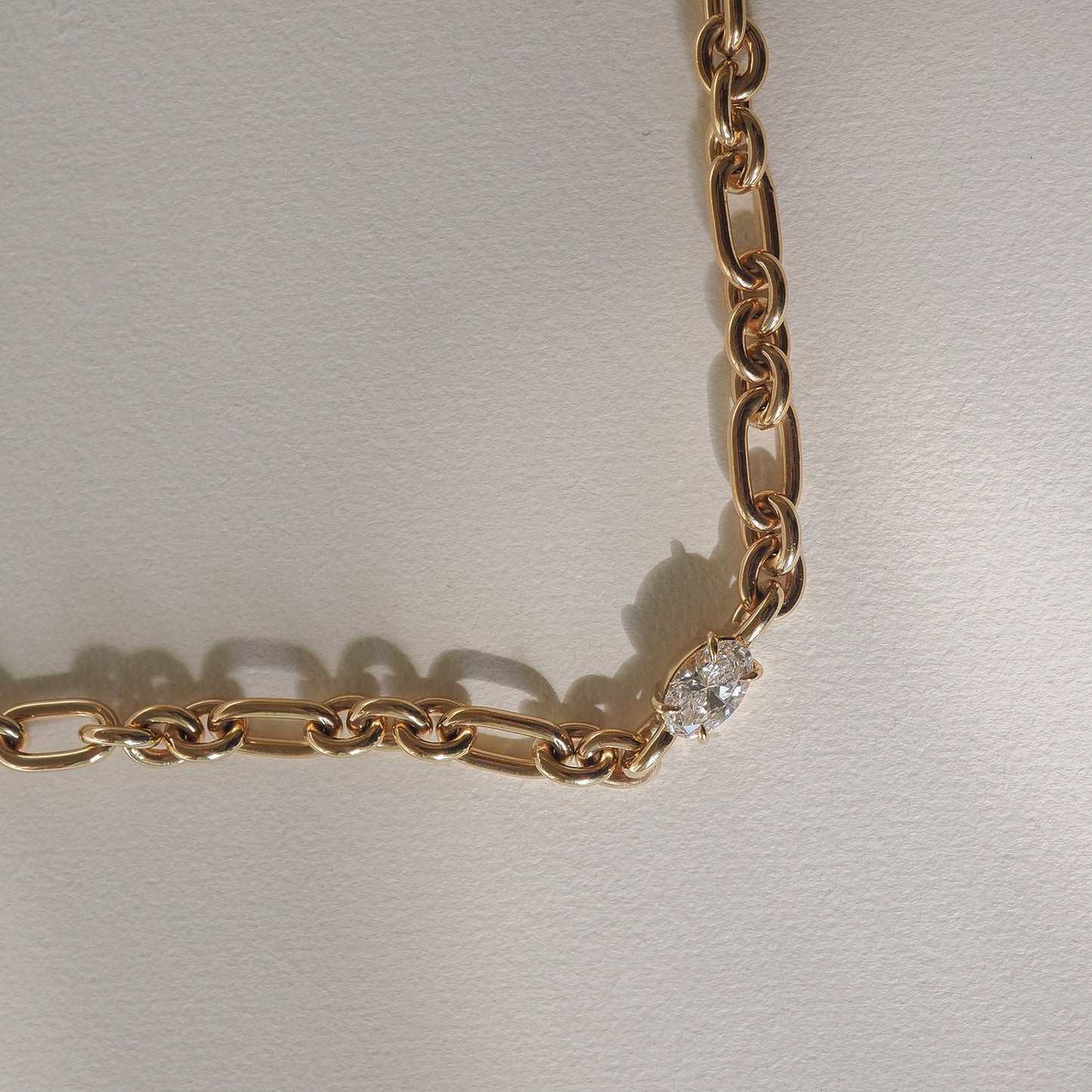 Gold chain necklace with a single sparkling diamond link, casting soft shadows on a cream surface. Luxurious jewellery design.