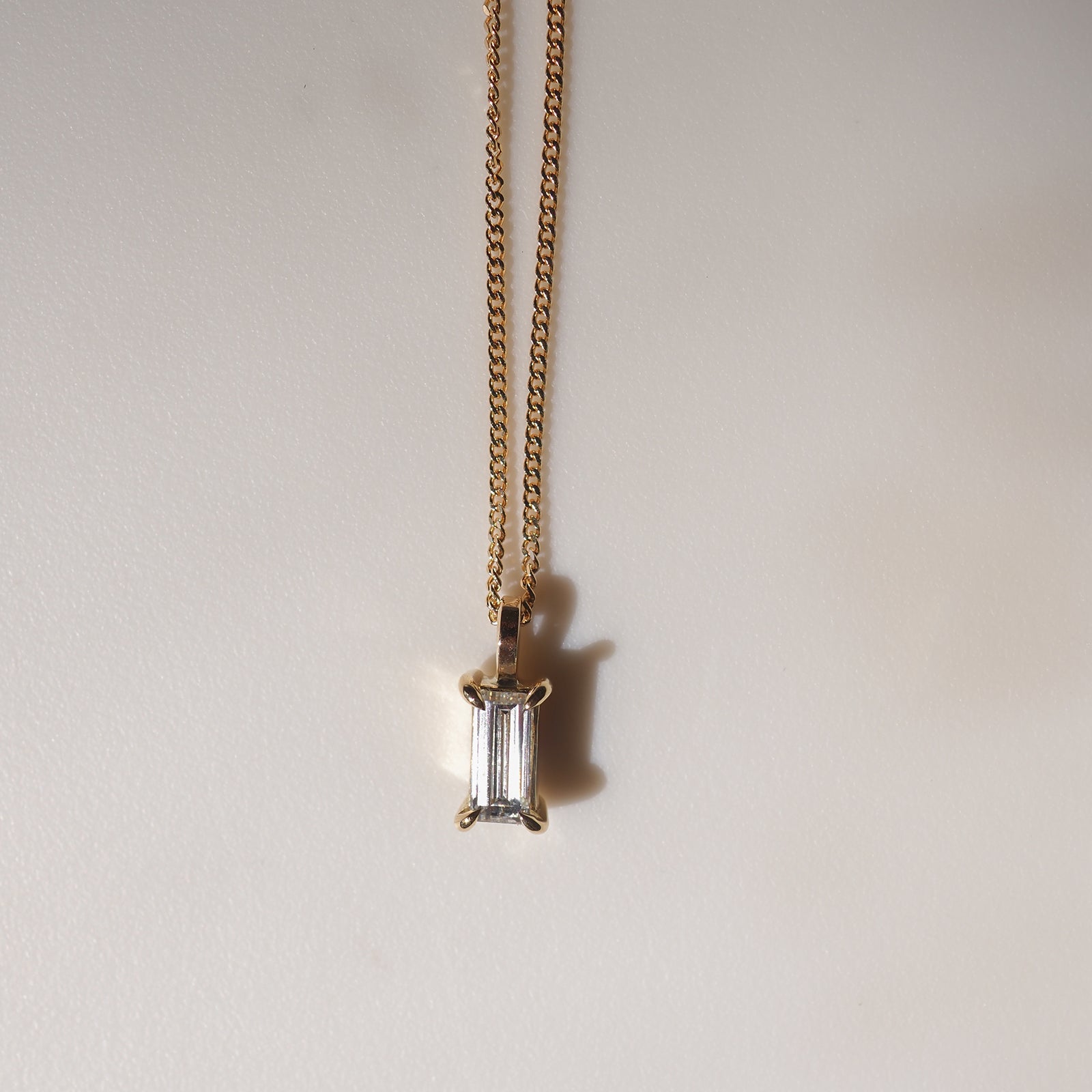 Gold chain necklace with an emerald-cut diamond pendant against a plain background, casting a subtle shadow. Luxurious and elegant design.