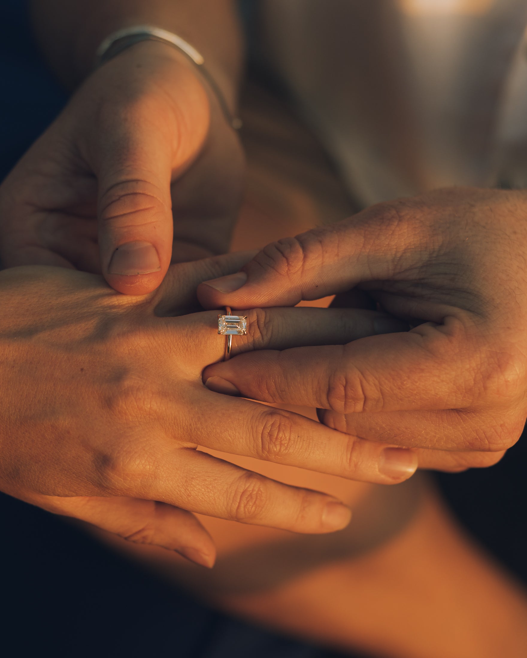 Close-up of hands exchanging an emerald-cut diamond engagement ring, emphasising luxury and elegance in soft, warm lighting.