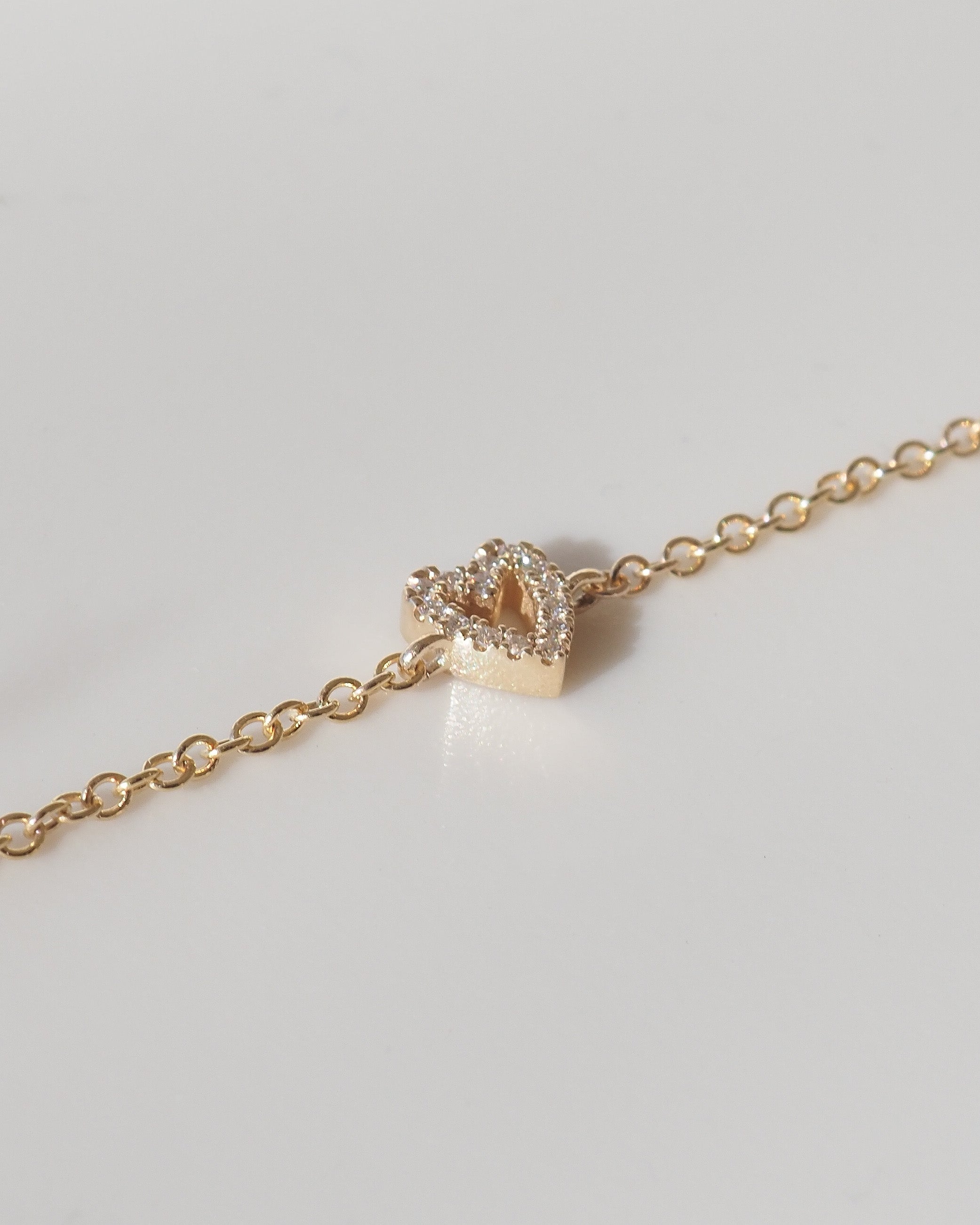 A delicate gold chain bracelet featuring a small, open heart-shaped charm encrusted with clear stones, displayed on a clean light gray background.