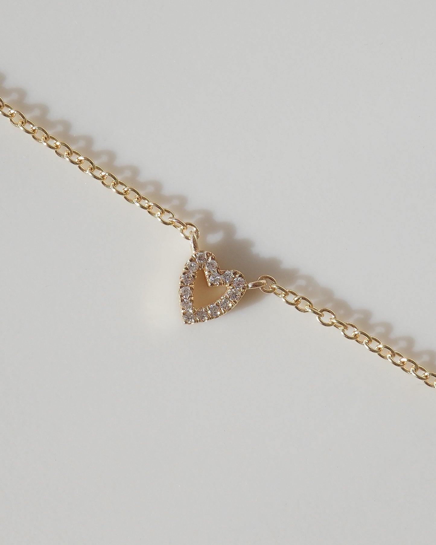 A delicate gold chain necklace featuring a small, open heart-shaped pendant encrusted with clear stones, displayed on a light gray background.
