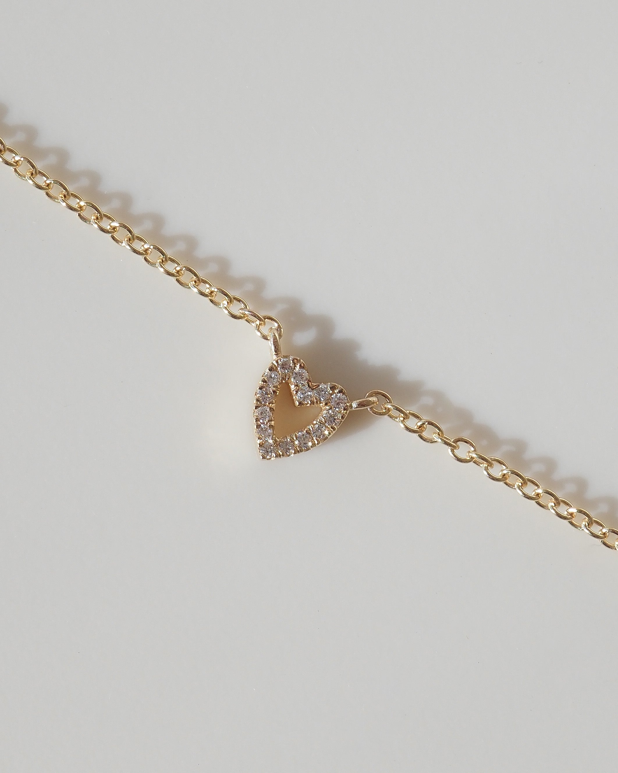 A delicate gold chain necklace featuring a small, open heart-shaped pendant encrusted with clear stones, displayed on a light gray background.