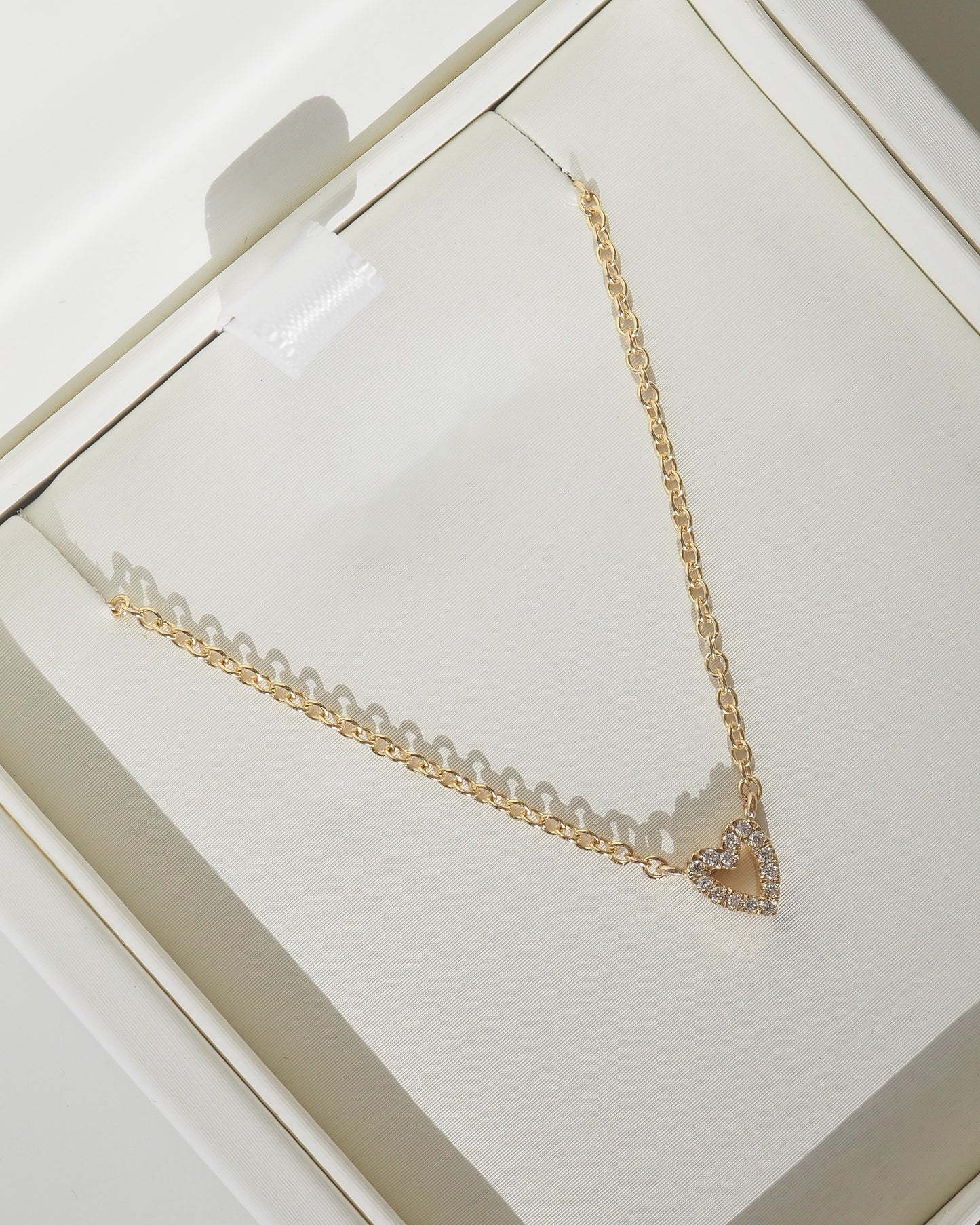 A gold chain necklace with a small heart-shaped pendant paved with clear stones, displayed inside a cream-colored jewelry box.
