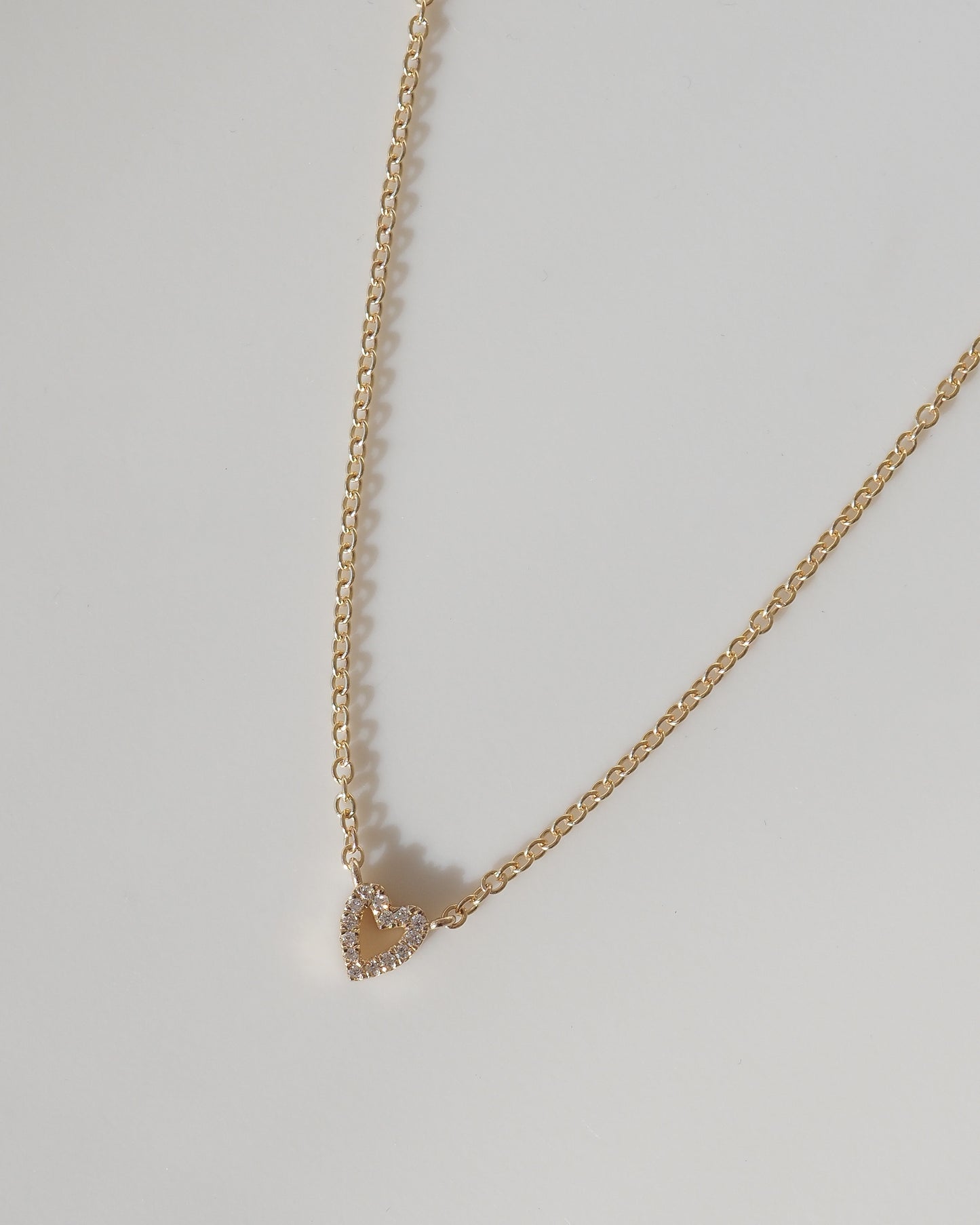A delicate gold chain necklace with a small, openwork heart-shaped pendant studded with tiny clear crystals, displayed on a neutral light gray background.