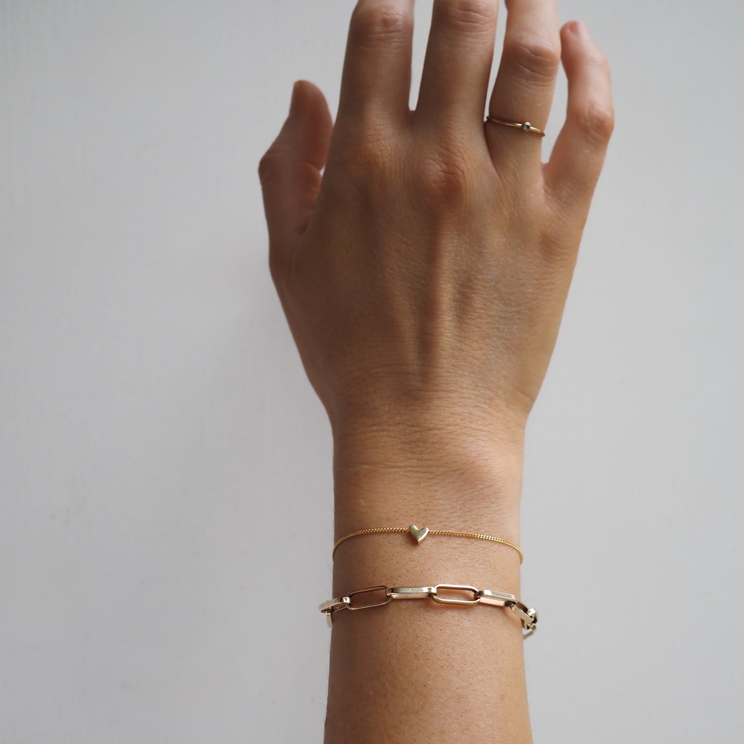 Hand wearing delicate gold bracelets and a gold ring. One bracelet features a small heart charm, and the other has chain links.