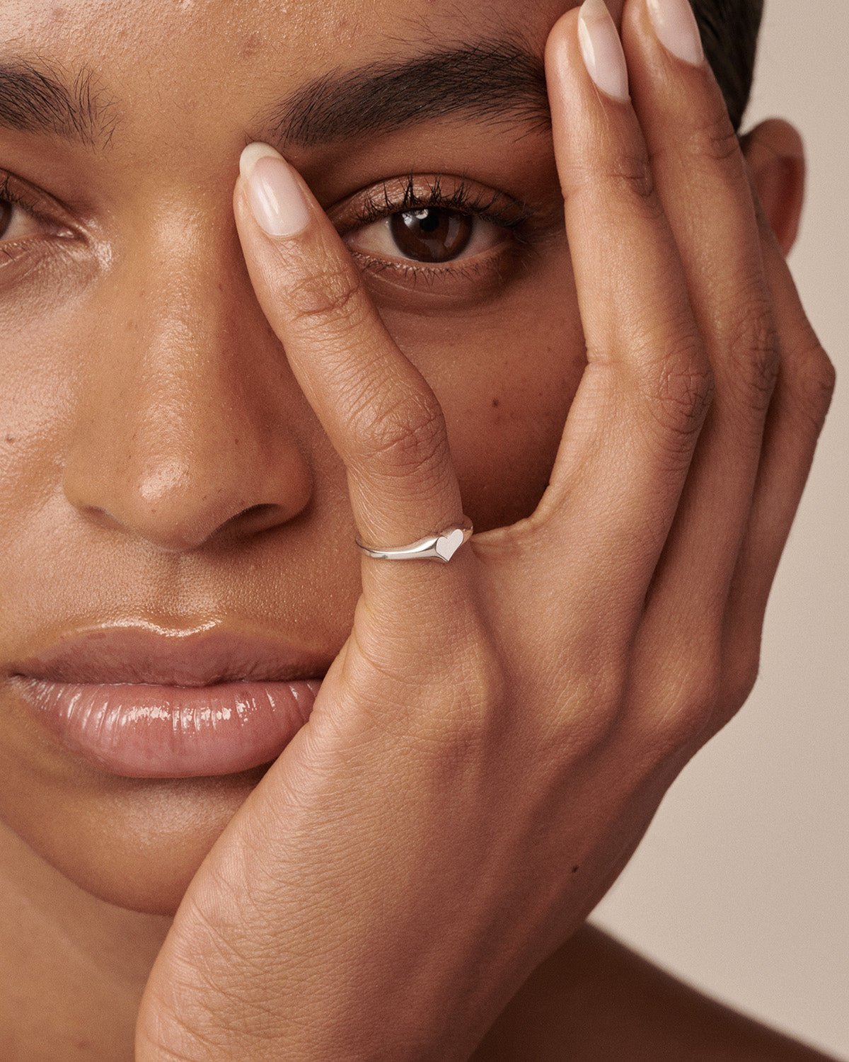 A close-up lifestyle shot of a model wearing a delicate silver band ring with a small, polished heart face on her finger, with her hand resting against her face.