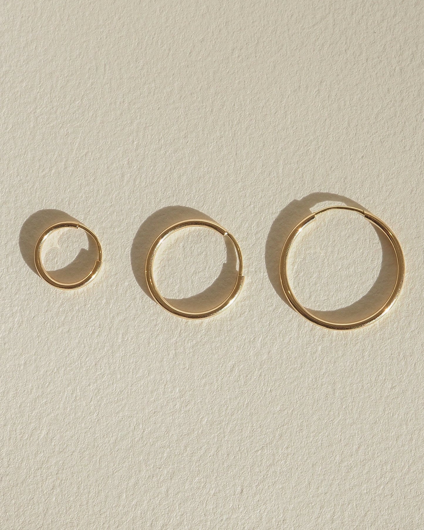 Four gold rings of varying sizes displayed on a textured beige surface, casting soft shadows. Elegant minimalist jewellery design.