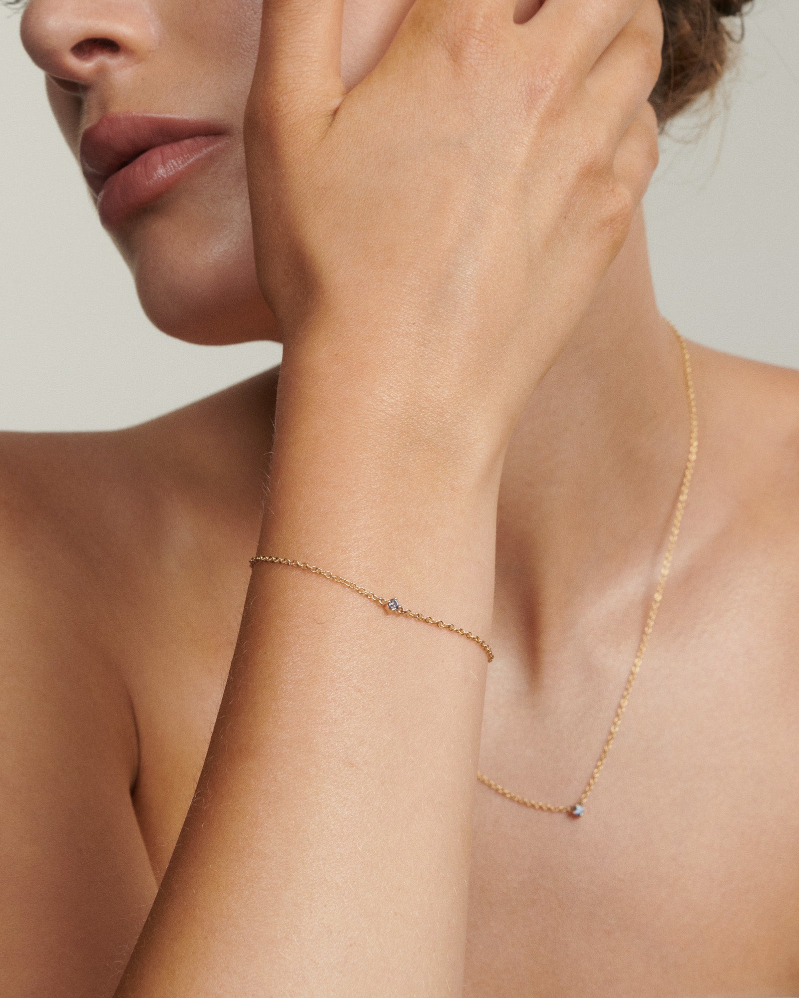 Close-up of a person wearing a delicate gold bracelet and necklace on a neutral background