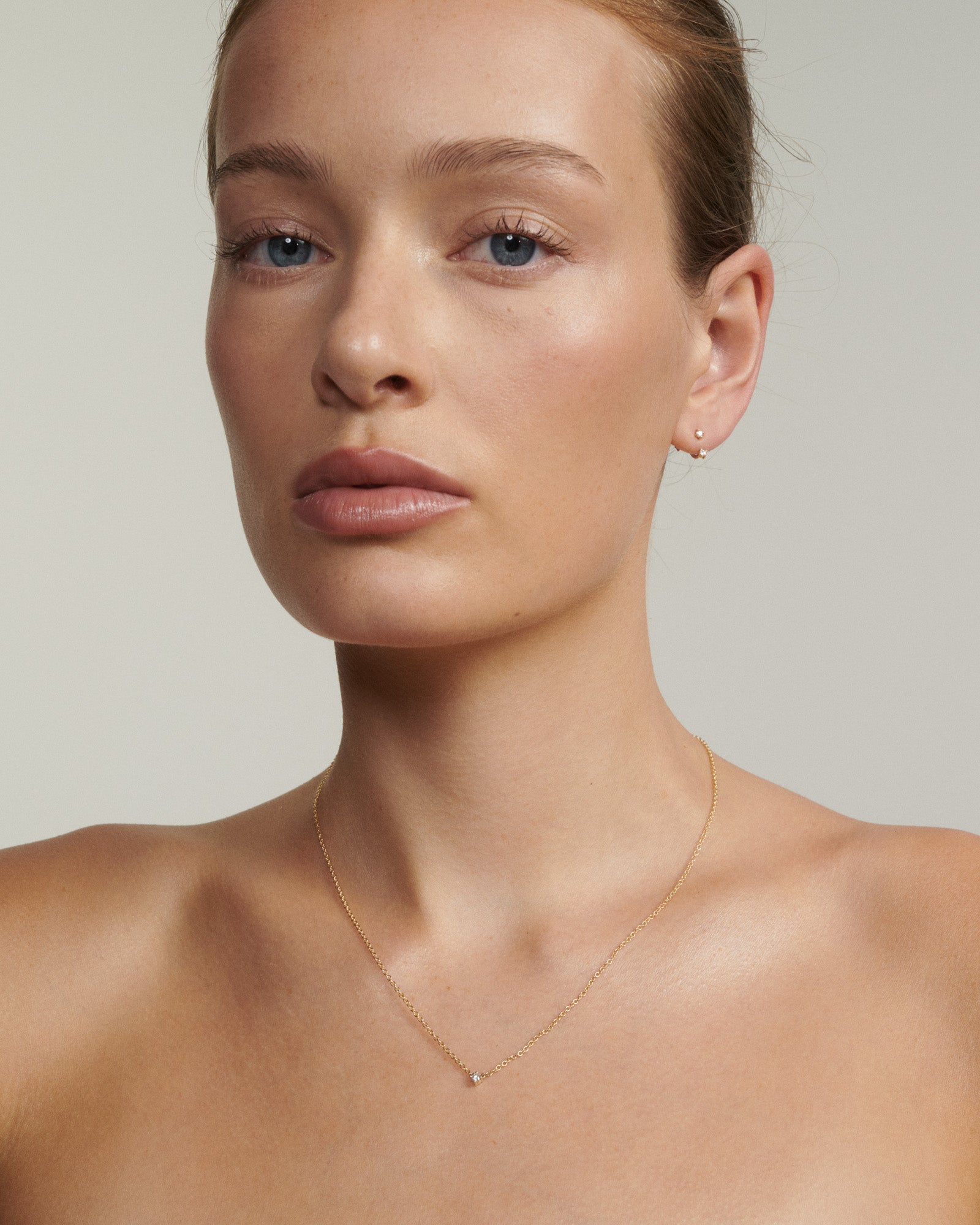 Close-up of a person wearing a delicate gold necklace and earrings against a neutral background [text overlay: Andrea wears the Tiny Birthstone Jackets, Studs and Necklace]