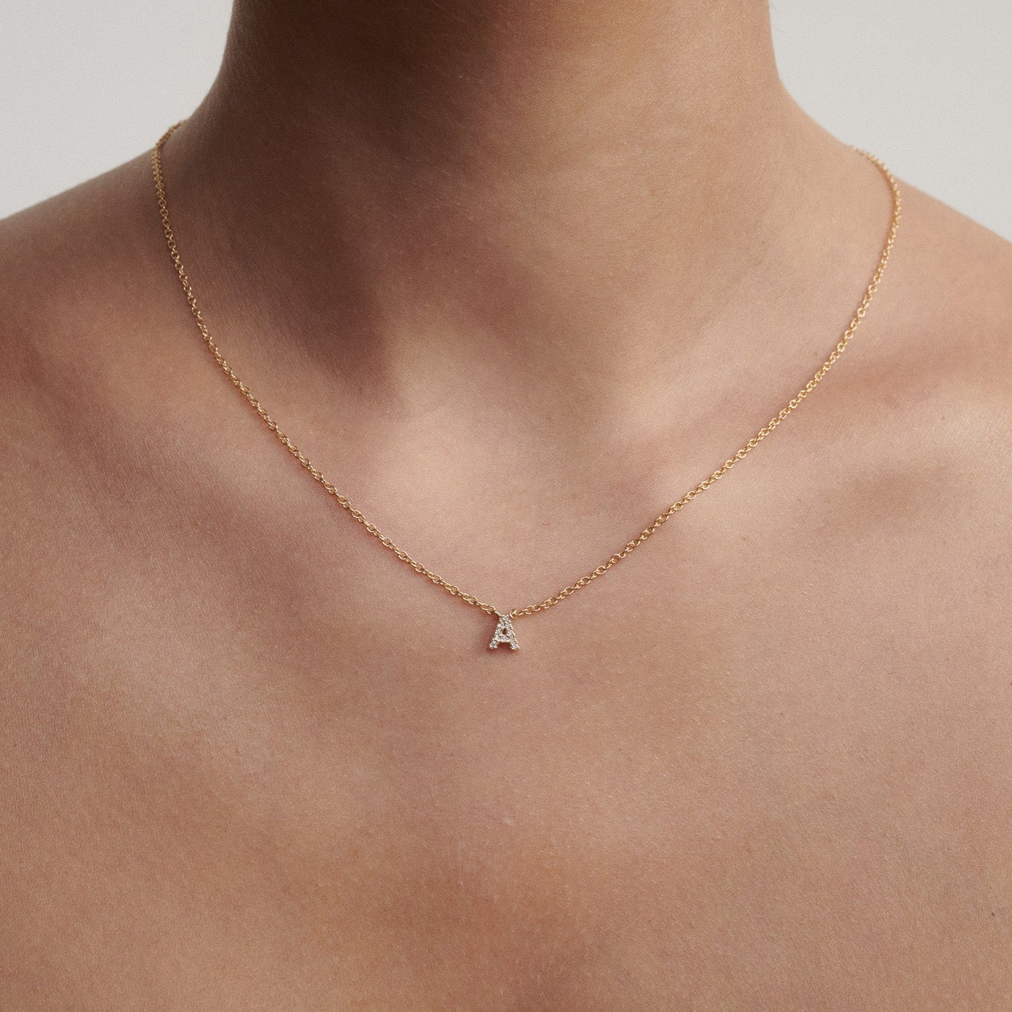 Gold necklace with a small diamond letter pendant on a person's neck against a neutral background