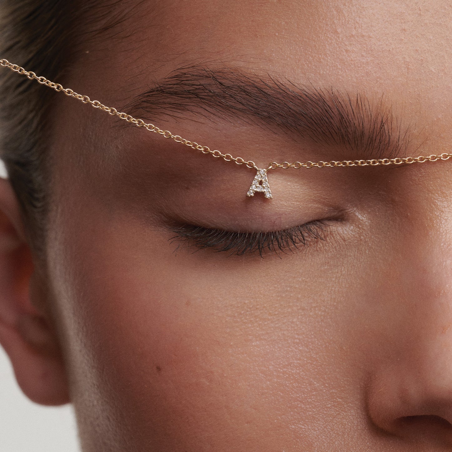 Close-up of a gold chain with a letter 'A' pendant worn across the eye in a creative way.