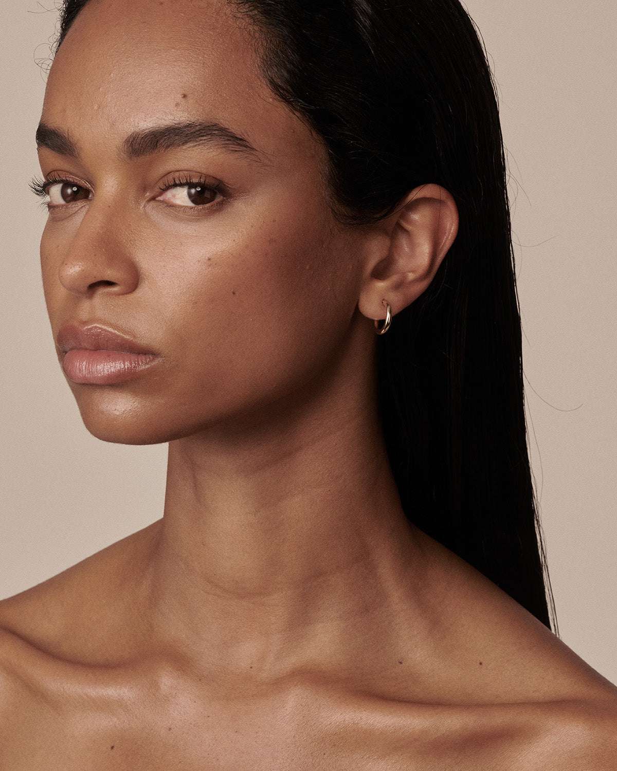 A close-up portrait of a woman with dark hair wearing a small gold hoop earring, set against a solid warm beige background. [text overlay: Francielle wears the 10mm Classic Hoops]