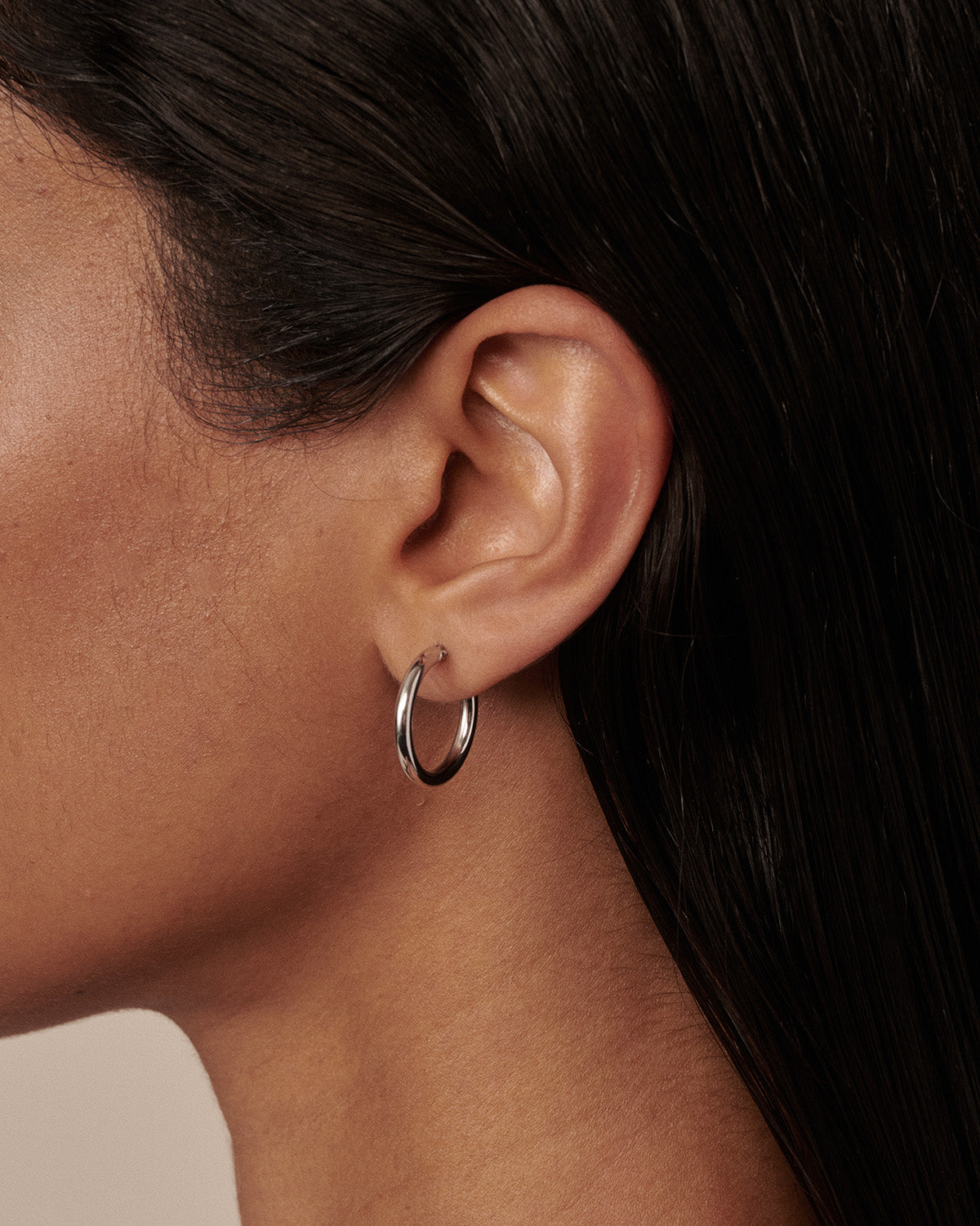 A close-up side profile of a model wearing a thin, polished silver hoop earring in her earlobe. [text overlay: Francielle wears the 15mm Classic Hoops]