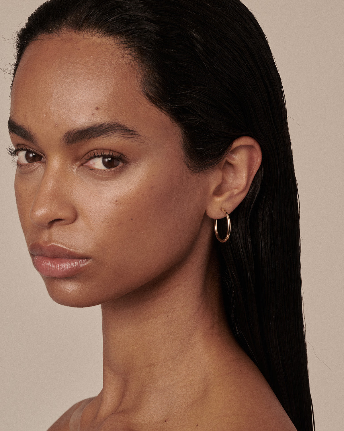 A close-up portrait of a woman with dark hair wearing a delicate gold hoop earring, set against a solid beige background. [text overlay: Francielle wears the 20mm Classic Hoops]