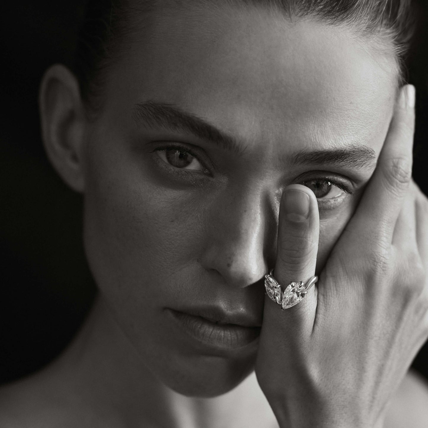 Black and white portrait of a person with a diamond ring on their finger, touching their face.