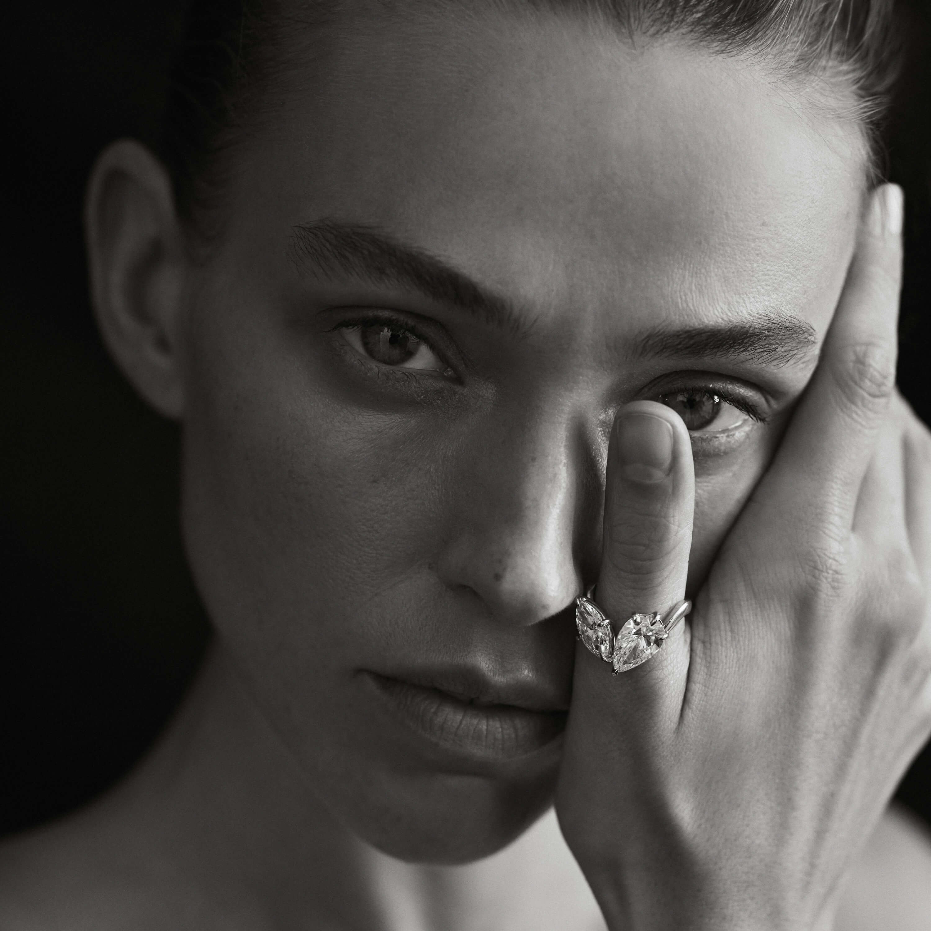 Black and white portrait of a person with a diamond ring on their finger, touching their face.
