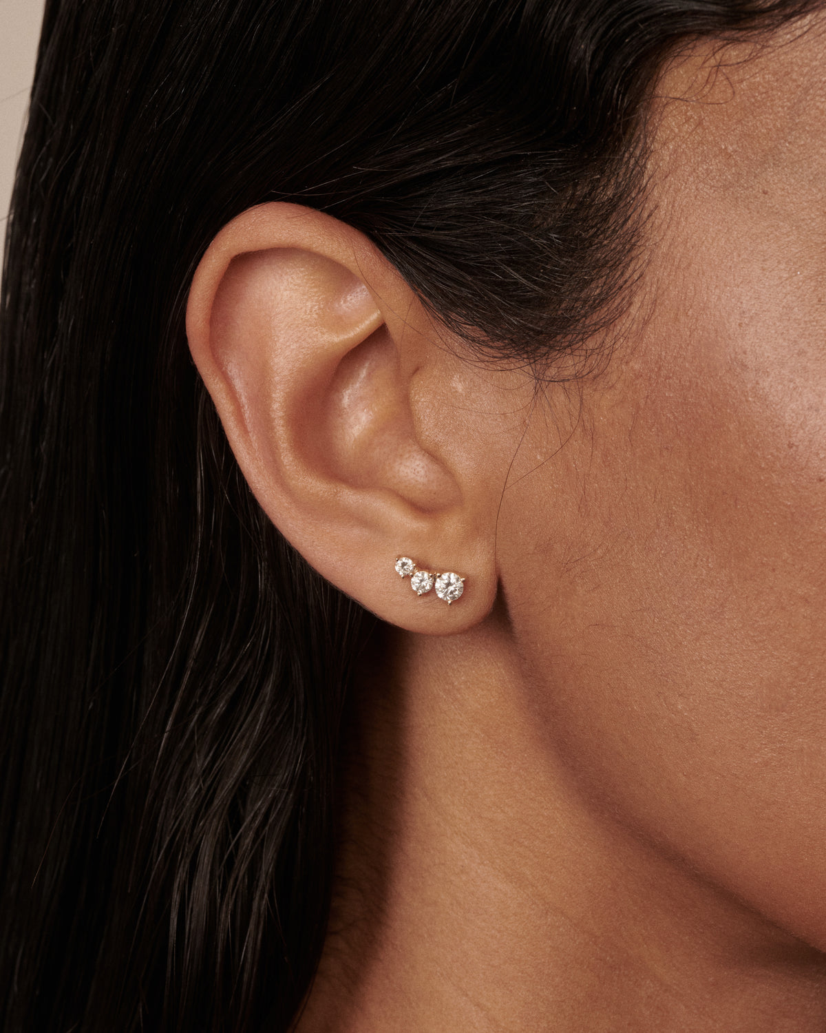 A close-up of a person's ear wearing a small cluster stud earring with three clear diamonds in a yellow gold setting.