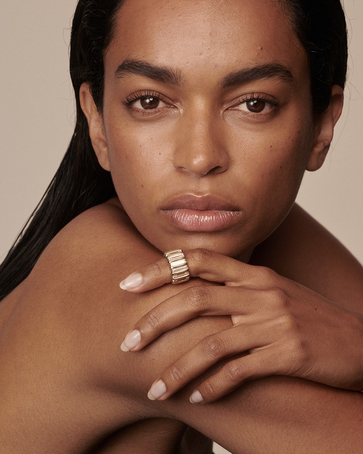 A close-up portrait of a model wearing a wide, textured gold ring on her finger, posed against a solid warm beige background. 