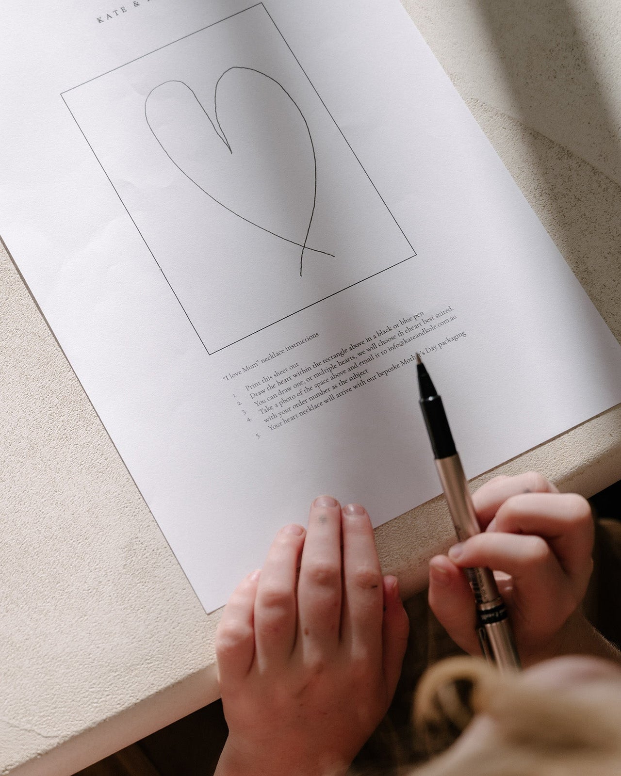 Hands hold a pen above a heart sketch on paper, suggesting luxury jewellery design. Elegant setting with soft lighting, evoking creativity.