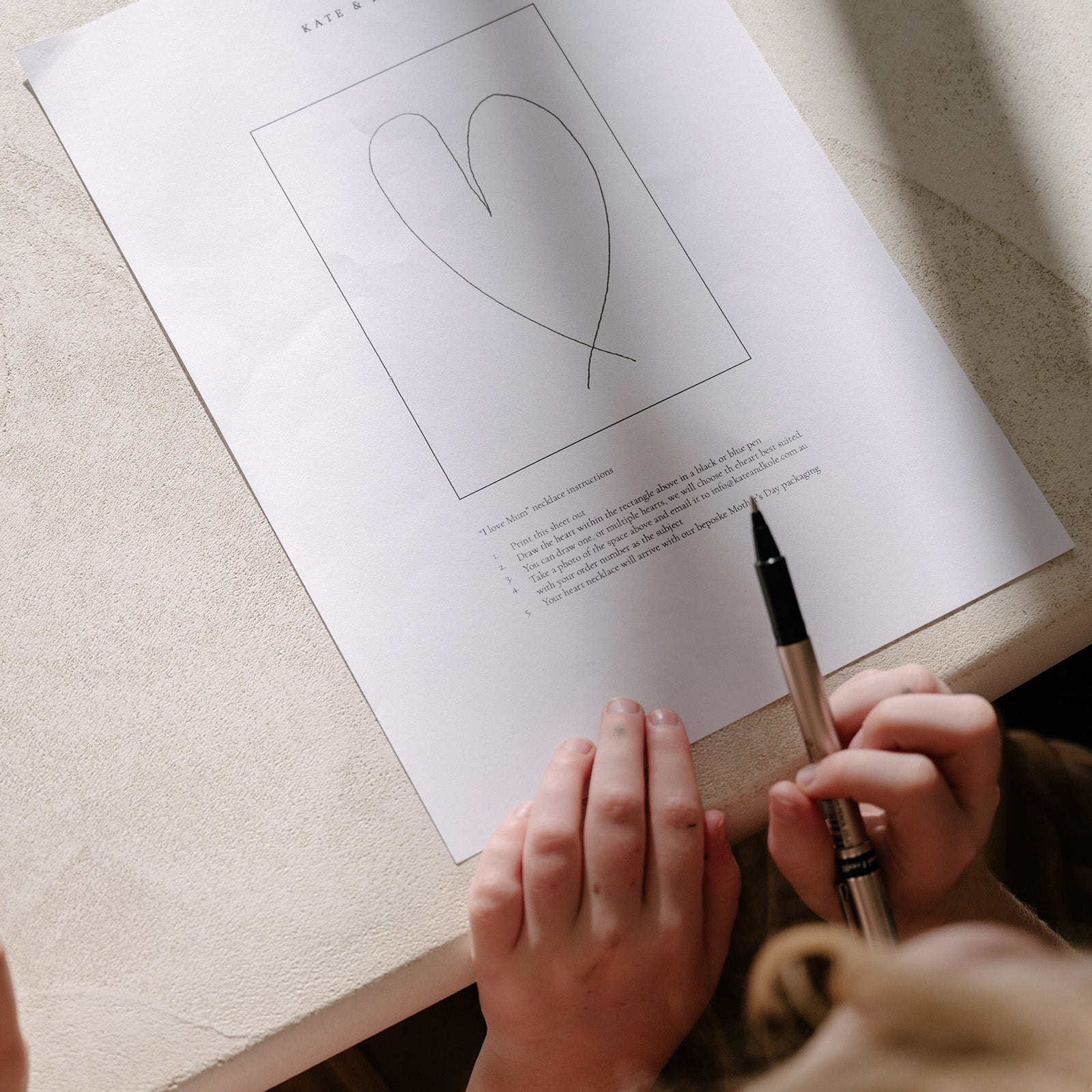 Hands hold a pen above a heart sketch on paper, suggesting luxury jewellery design. Elegant setting with soft lighting, evoking creativity.