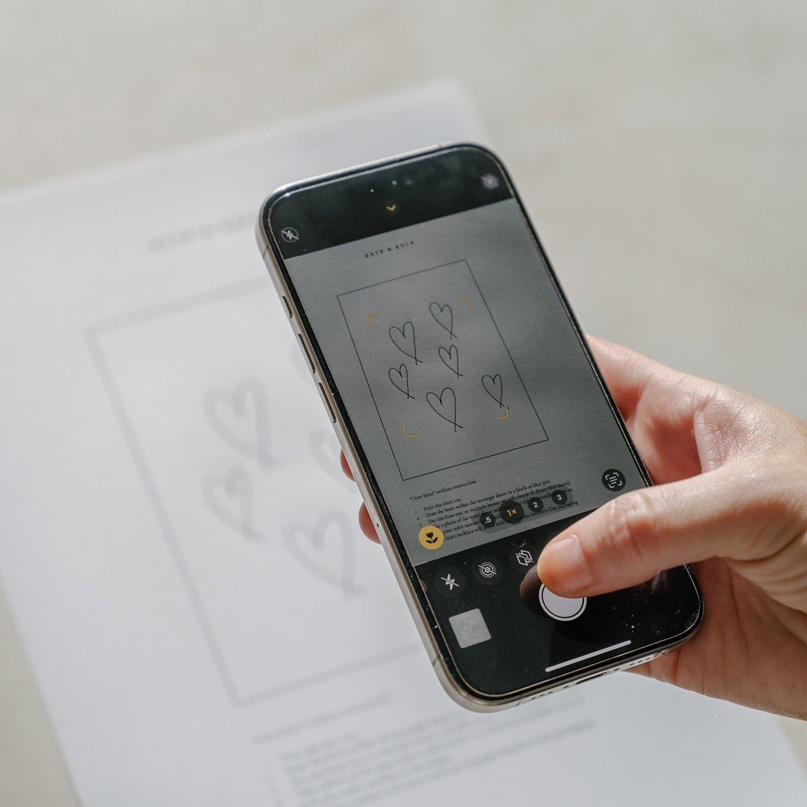 Hand holding a smartphone, capturing a photo of a paper with heart designs, possibly related to luxury jewellery sketches.