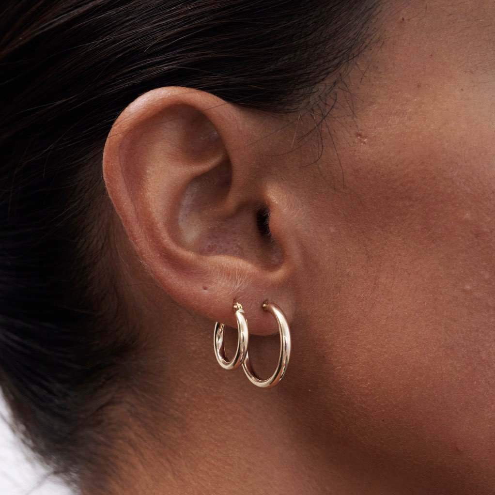 Close-up of a woman's ear wearing two sleek gold hoops, highlighting luxury jewellery fashion.[text overlay: 10mm Hoop (left) & 15mm Hoop (right)] 