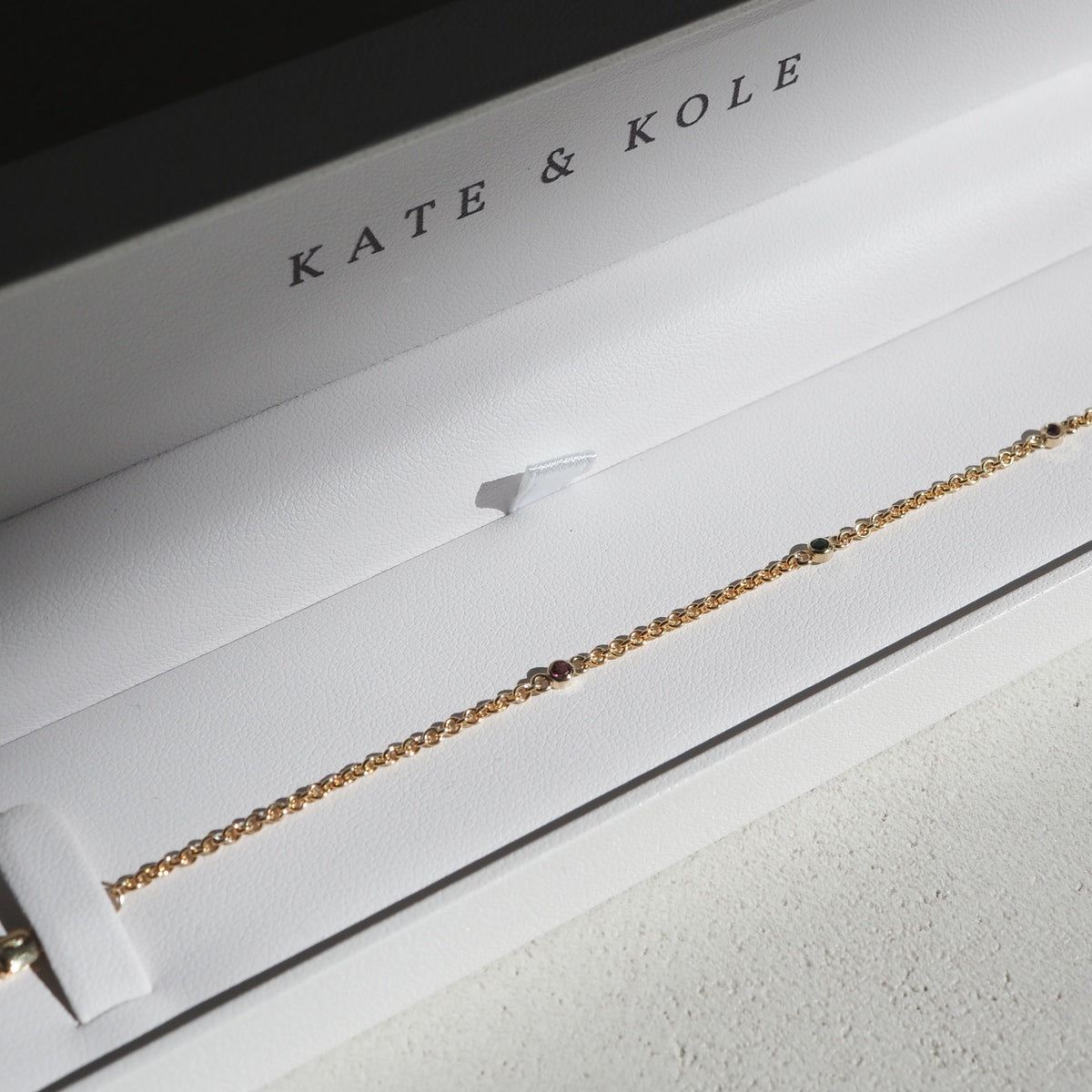 Gold chain bracelet with gems in a white Kate & Kole box, set on a textured surface. Luxurious jewellery, elegant design.