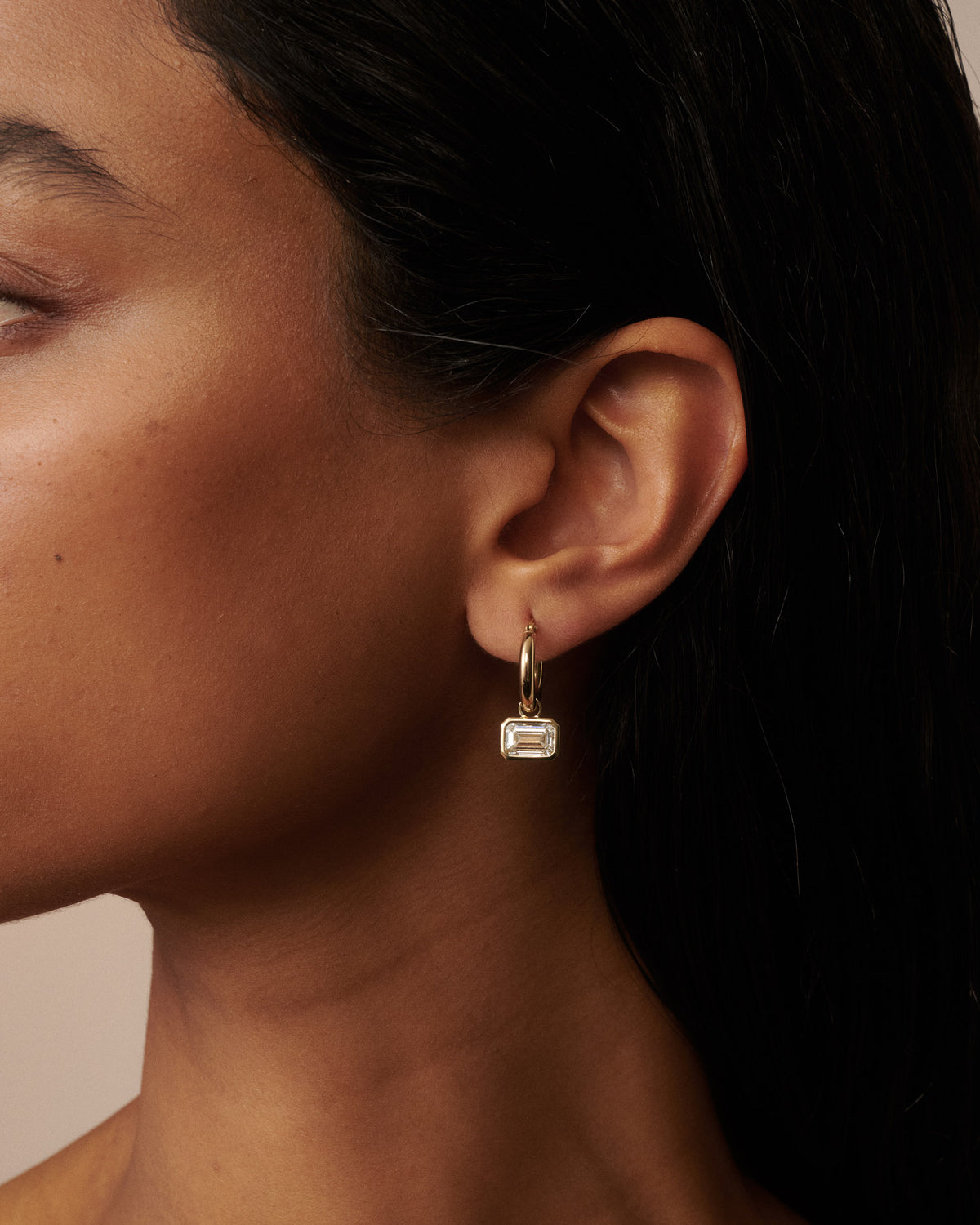 A close-up of a model's ear wearing a delicate gold huggie hoop earring with a small rectangular clear gemstone  dangle.