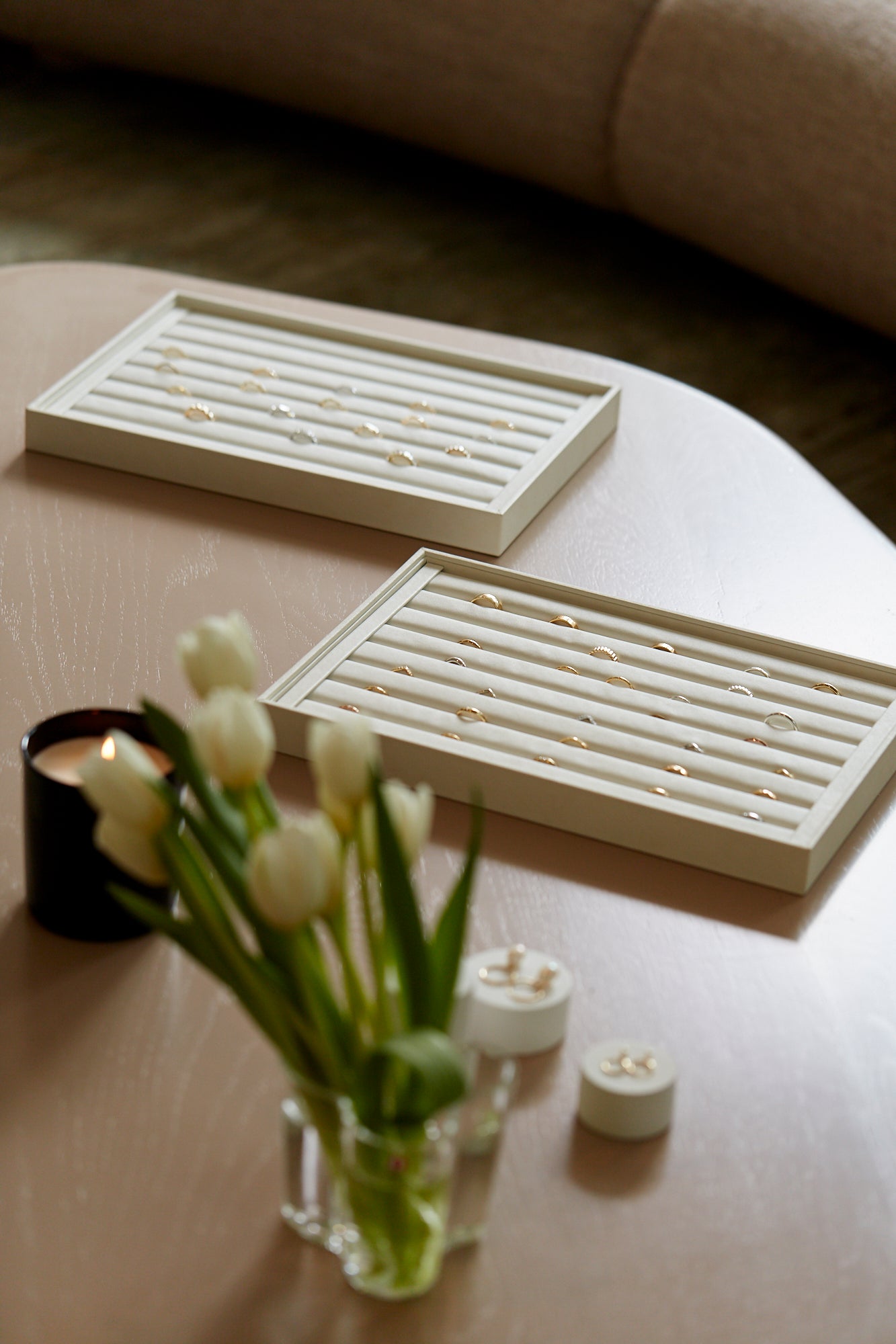 Luxury jewellery display with gold rings on cream trays on a wooden table. Tulips in a glass vase and a lit candle add elegance.
