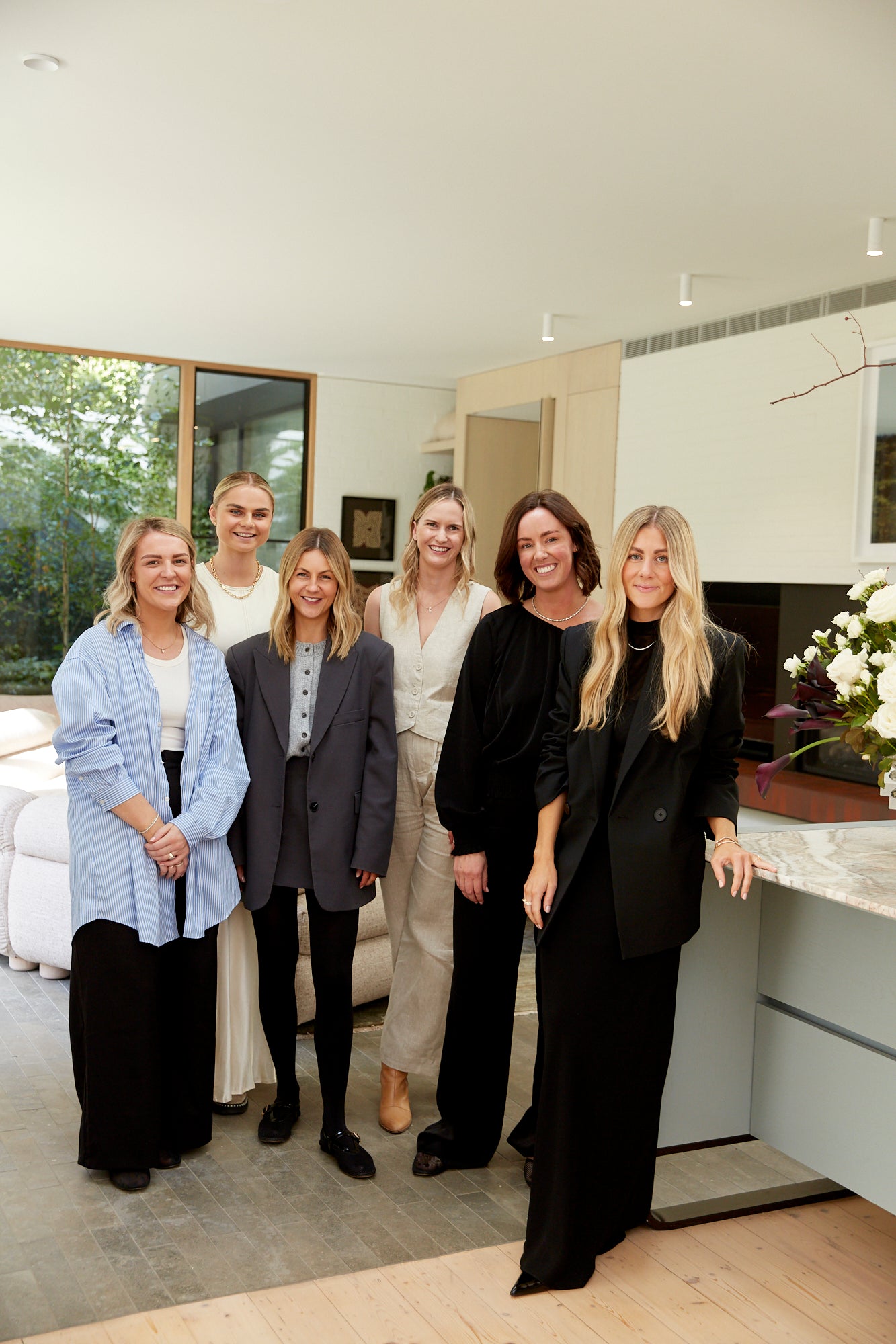 A group of six women in stylish, professional attire stand indoors with a modern living room and plants in the background.