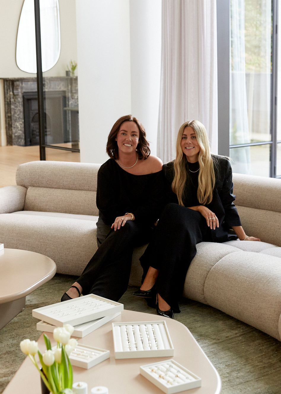Two women in black sit on a cream sofa in a modern, luxurious room with white tulips. Jewellery displays on the table add an elegant touch.