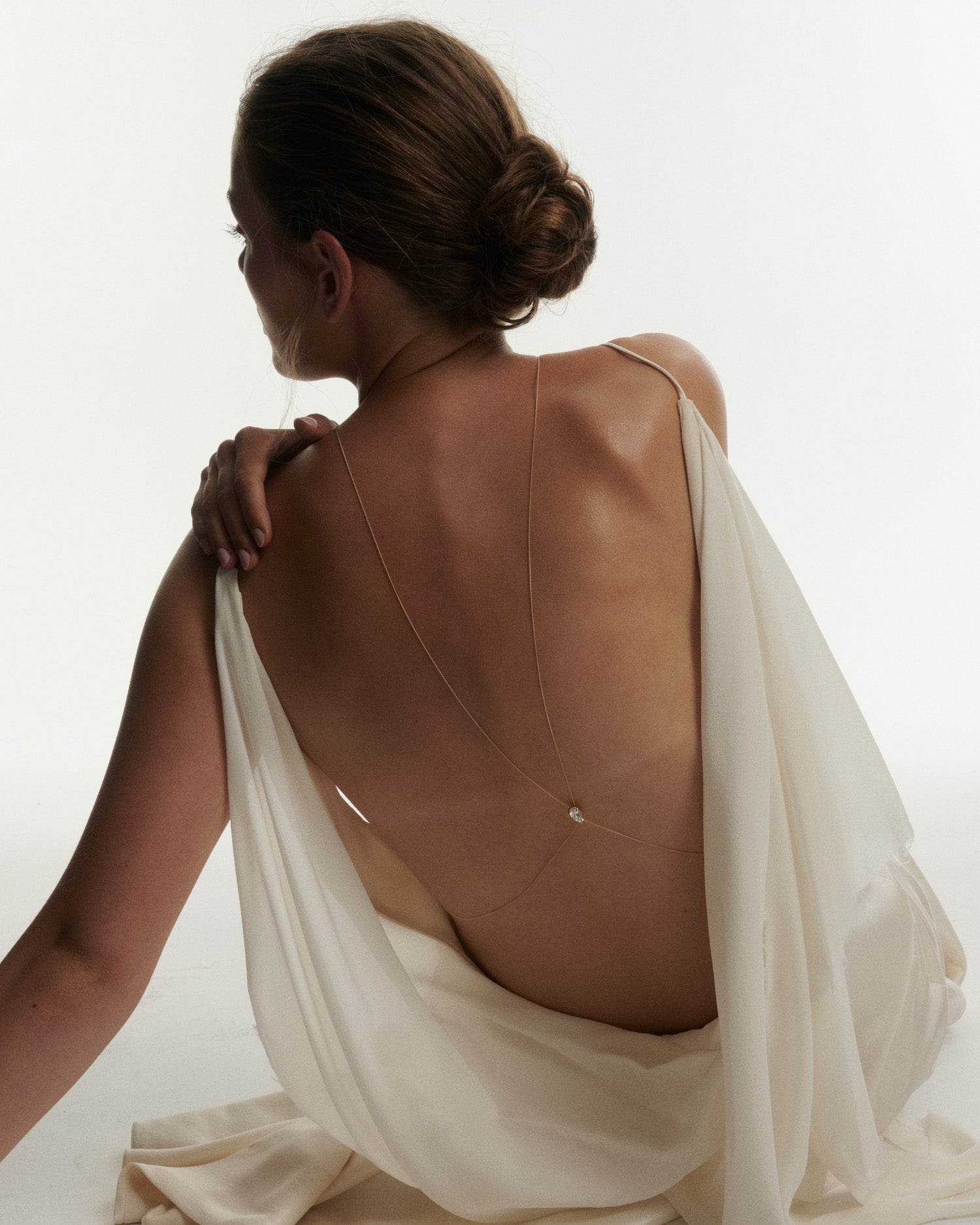 Woman wearing a beige leather backless dress with a white fabric draped over her shoulders on a plain background