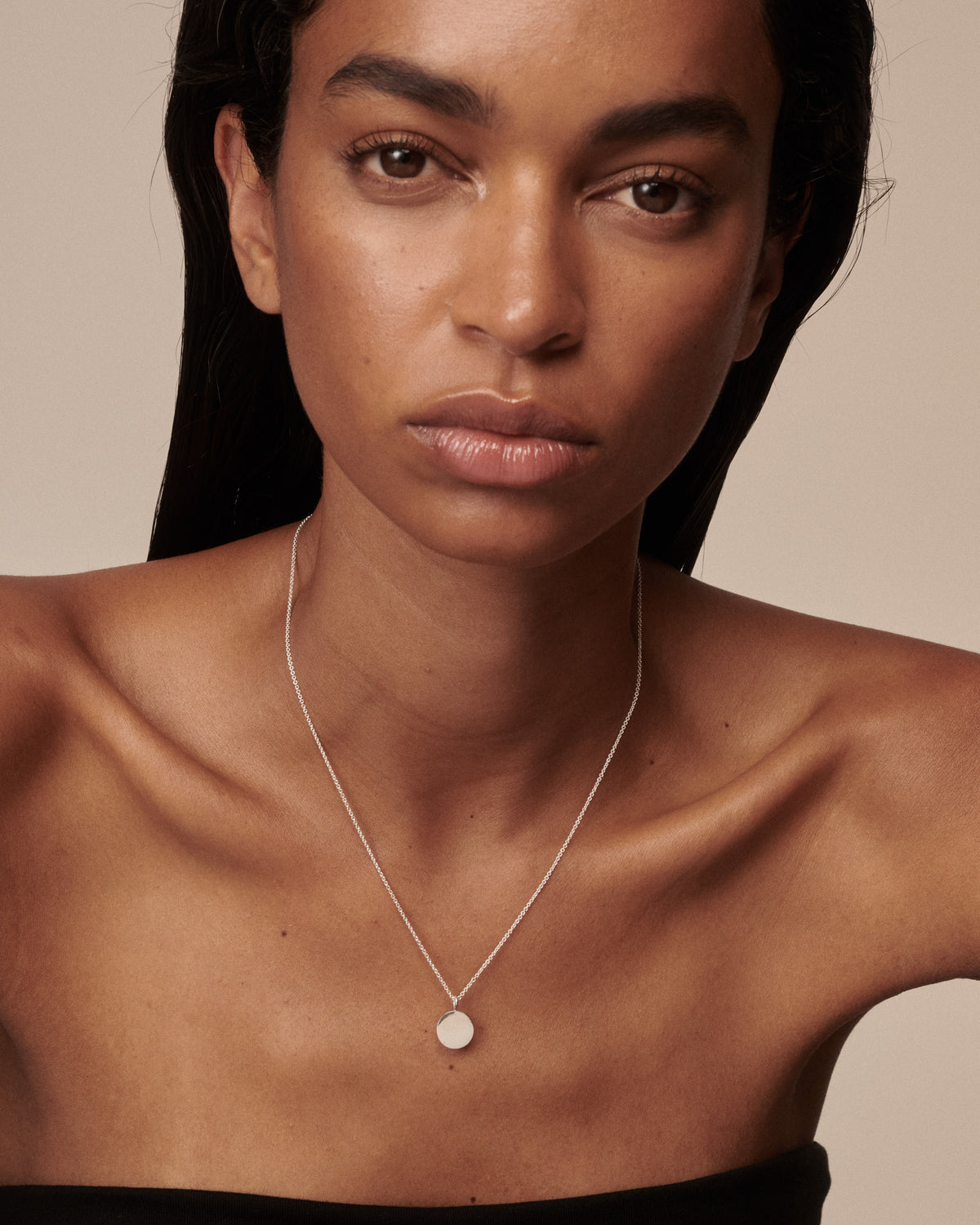A close-up of a model wearing a delicate silver chain necklace with a small, round, disc-shaped pendant on a warm beige background.