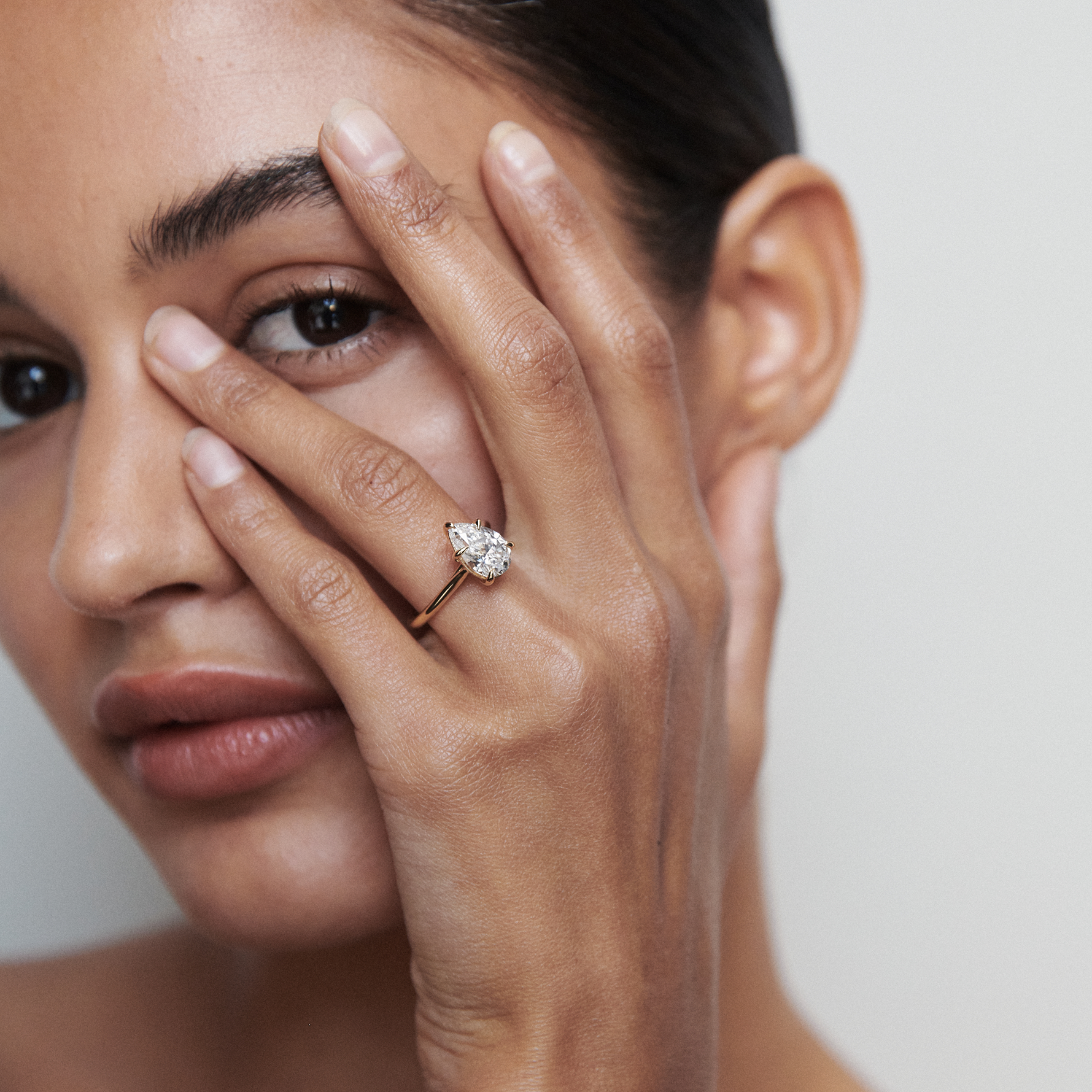 Woman with sleek bun poses, displaying a pear-shaped diamond ring on her finger, close to her face. Luxurious jewellery detail.