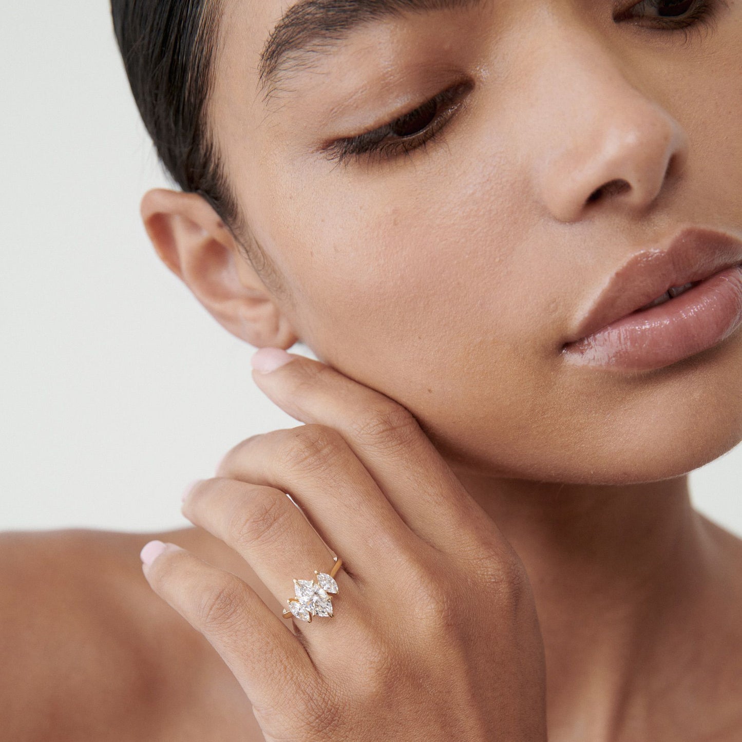 Woman with nude lips wears a large diamond cluster ring on her finger, showcasing luxury jewellery against a soft, neutral background.