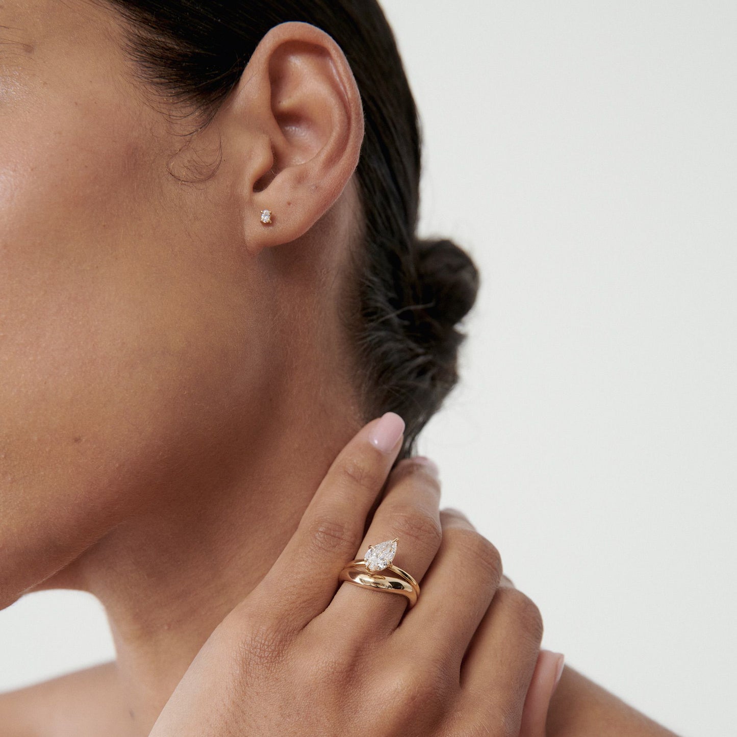 Close-up of a woman wearing a diamond stud earring and a delicate gold ring with a marquise diamond, against a neutral background.