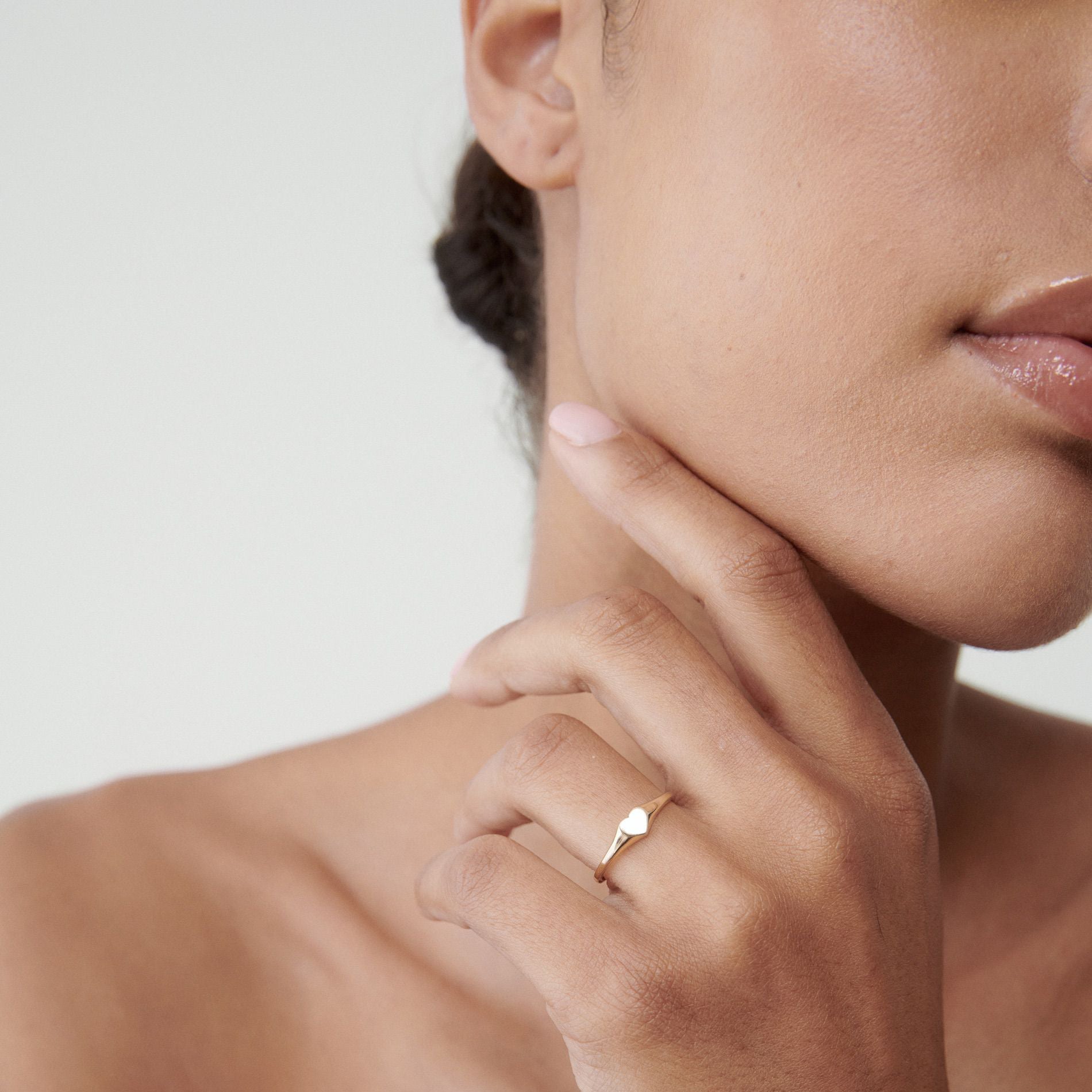 Woman's hand touching chin, wearing a delicate gold signet ring. Close-up on jewellery against soft skin tones. Elegant and understated luxury.