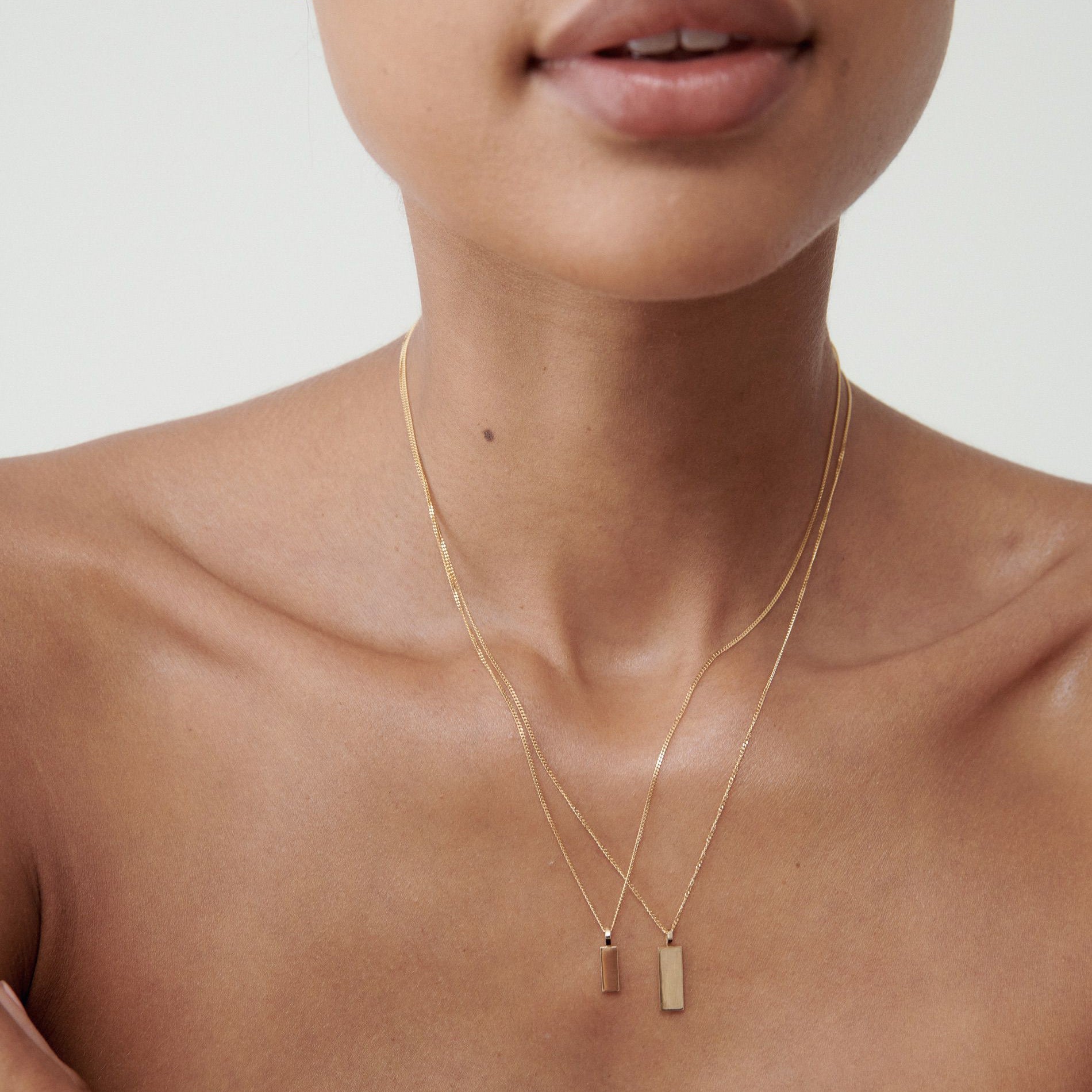 Close-up of a person wearing two delicate gold necklaces with rectangular pendants, featuring minimalistic jewellery design.