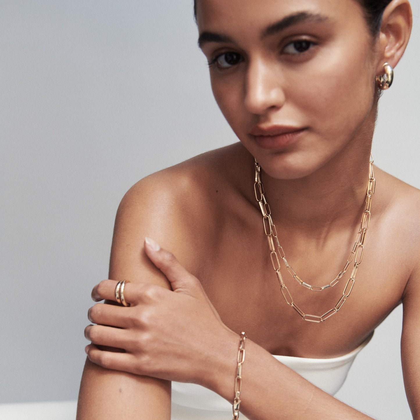 Woman wearing elegant gold jewellery, including hoop earrings, layered chain necklaces, rings, and bracelet. Luxurious style, soft background.