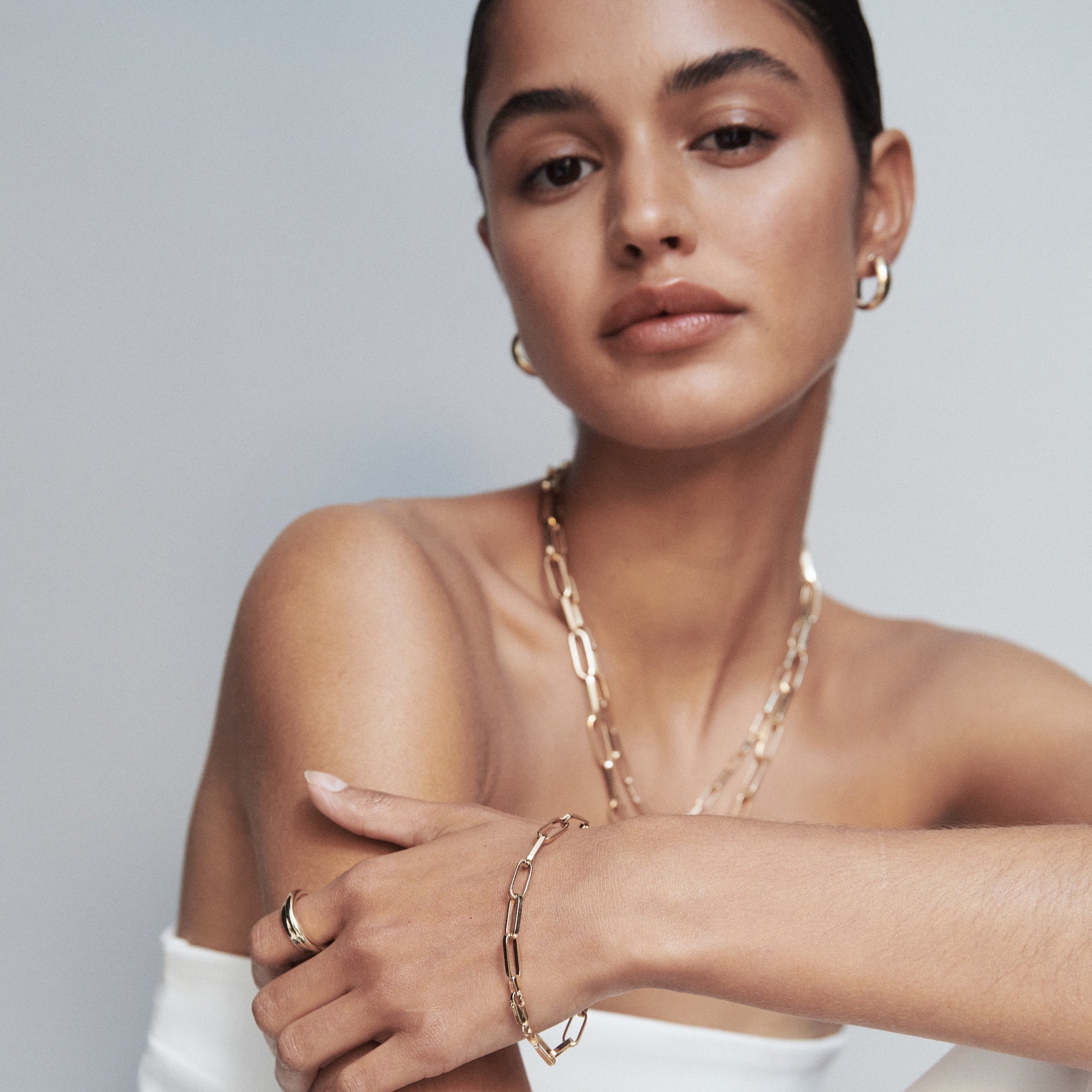 Woman wearing gold hoop earrings, layered chain necklaces, bracelet, and ring, against a neutral background. Luxe jewellery fashion.