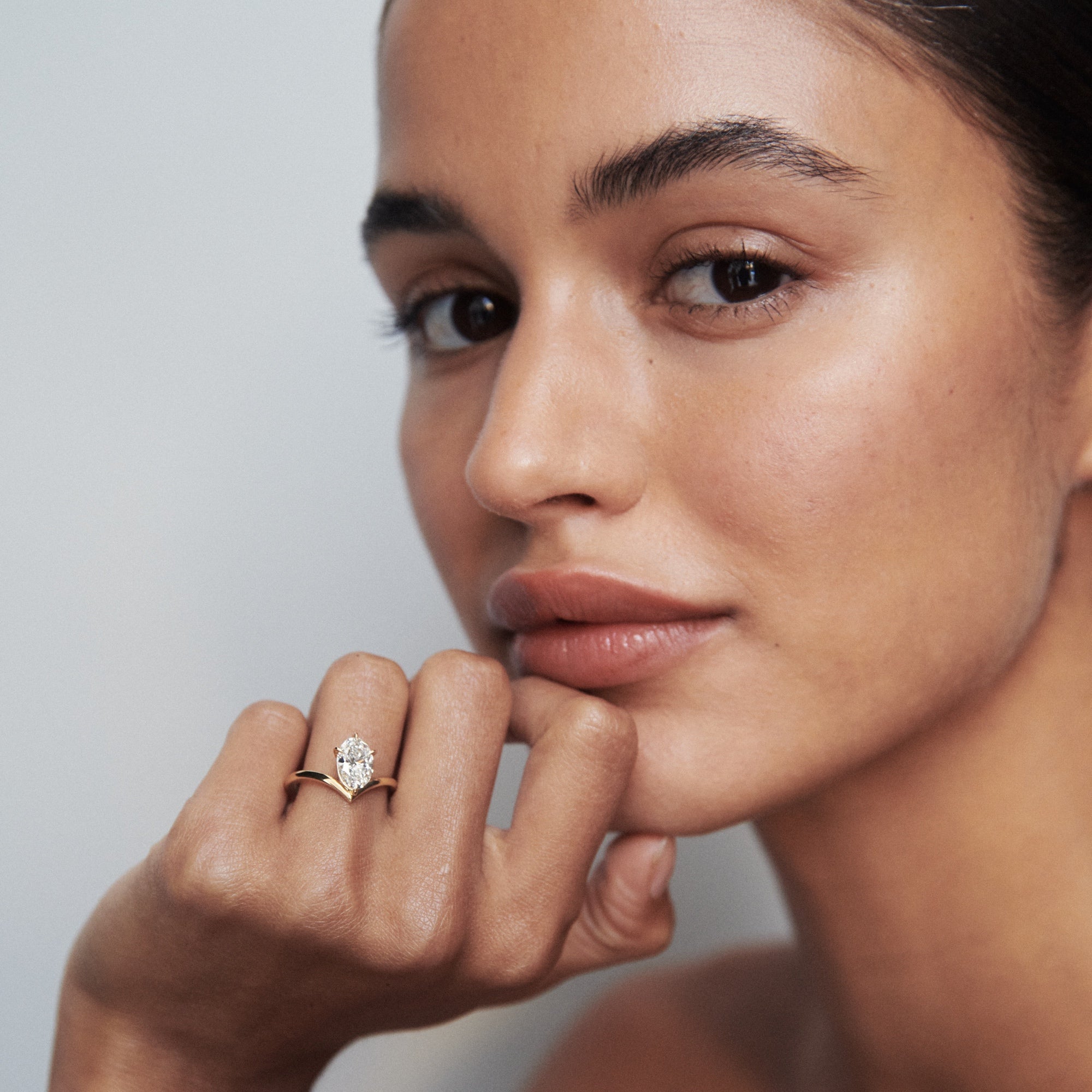 Woman showcasing a pear-cut diamond ring on her finger, resting her chin on her hand, with elegant, natural makeup and a serene expression.
