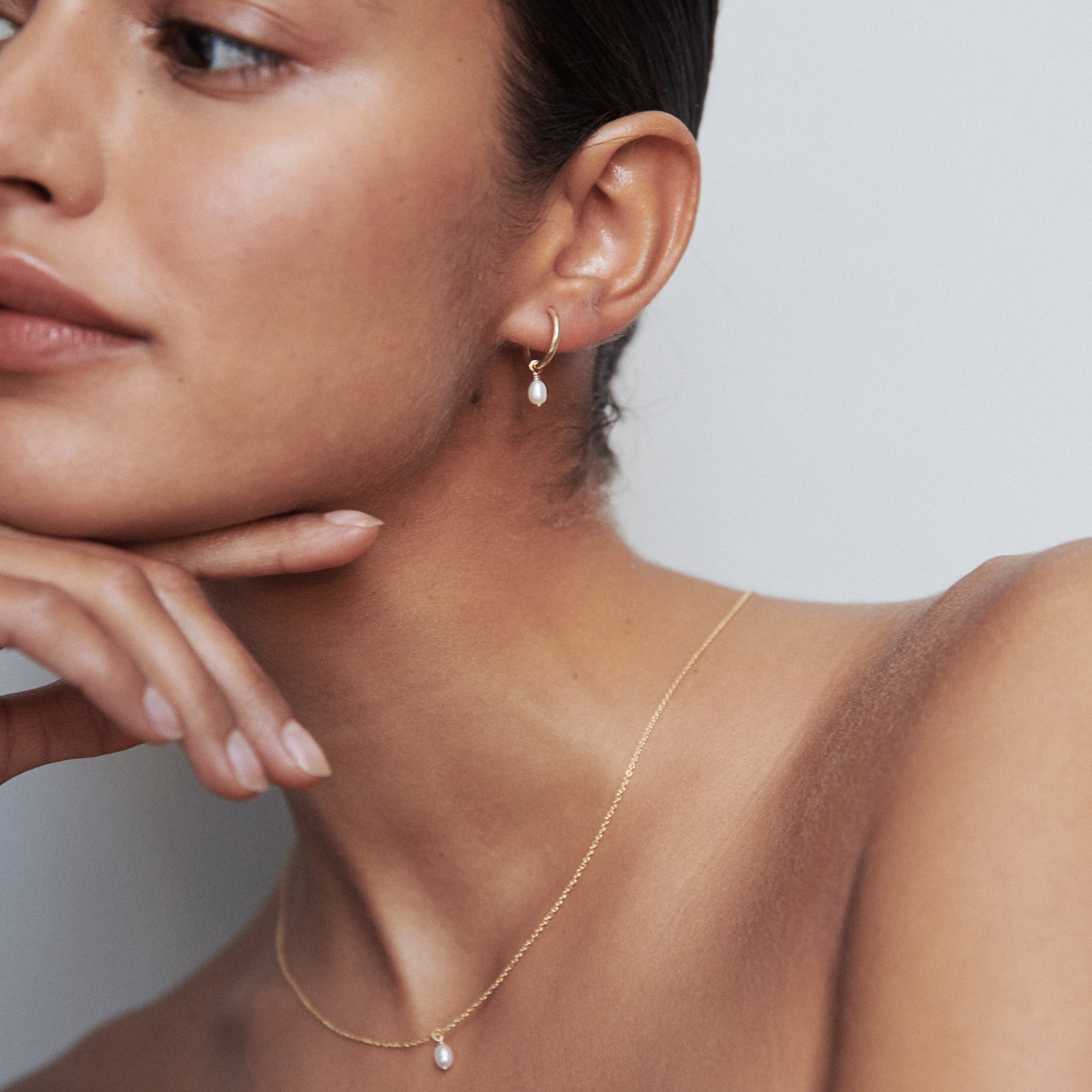 Woman wearing elegant gold hoop earrings and a fine gold necklace with a small pendant. Minimalist luxury jewellery.[text overlay: Tiny Pearl Necklace (Pictured)] 
