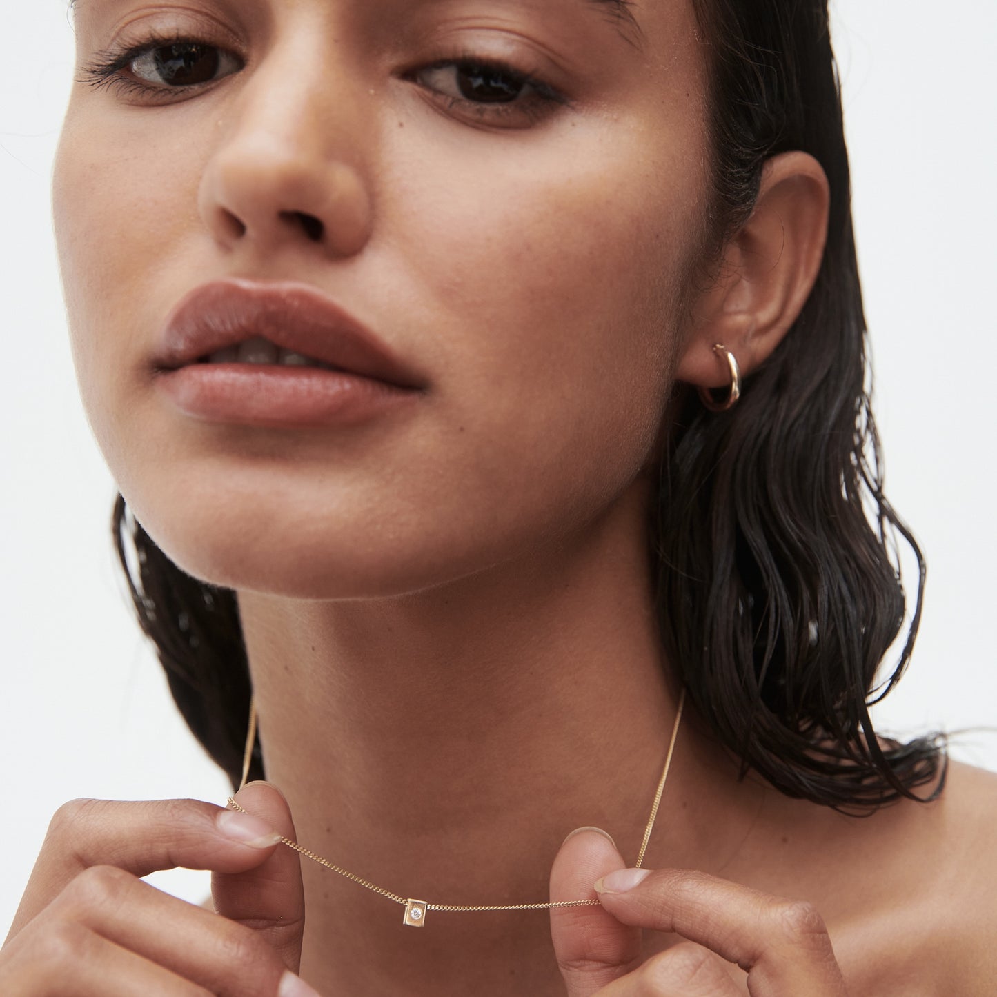 Woman with wet hair wears a delicate gold necklace featuring a small square pendant and a gold hoop earring, set against a white background.[text overlay: April Birthstone]
