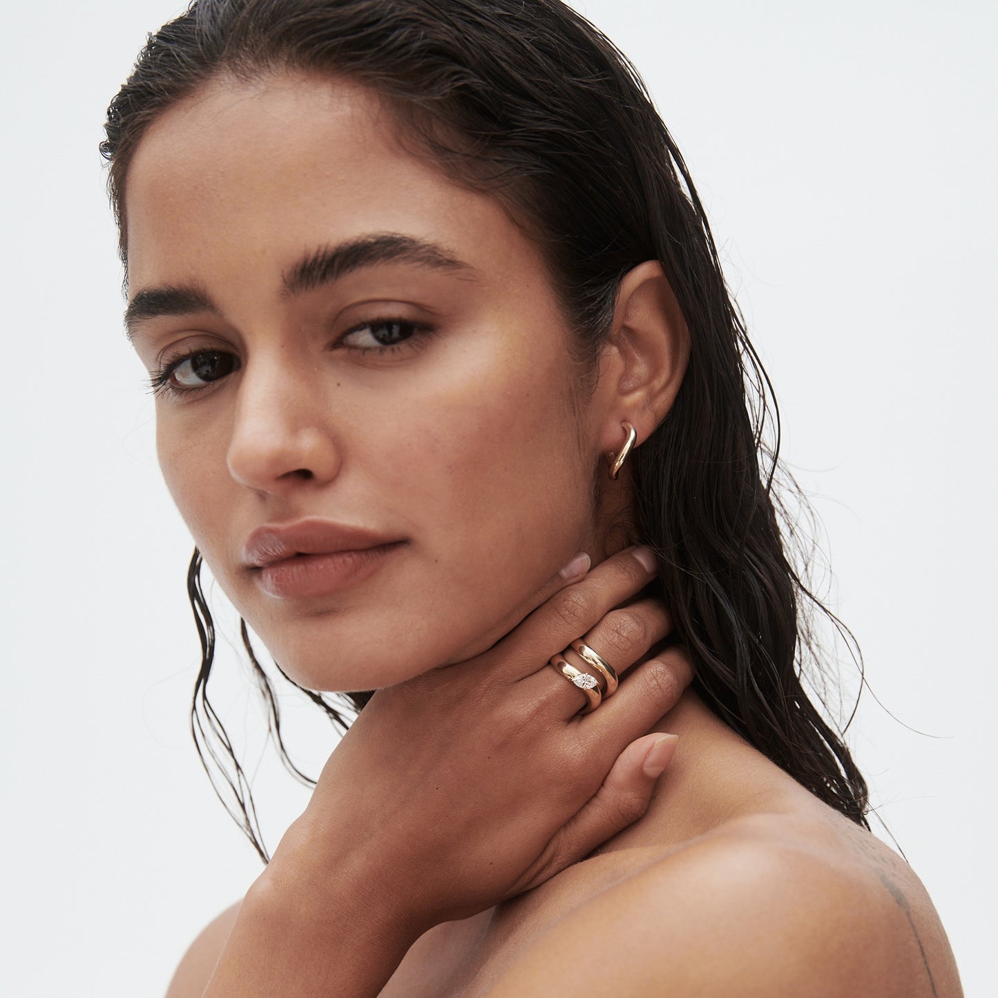 Woman with wet hair wears gold hoop earrings and stacked rings, hand on neck, against a plain background. Luxury jewellery portrait. [text overlay: Bold with Awa Bold Marquise Diamond Ring][text colour: #000000]
