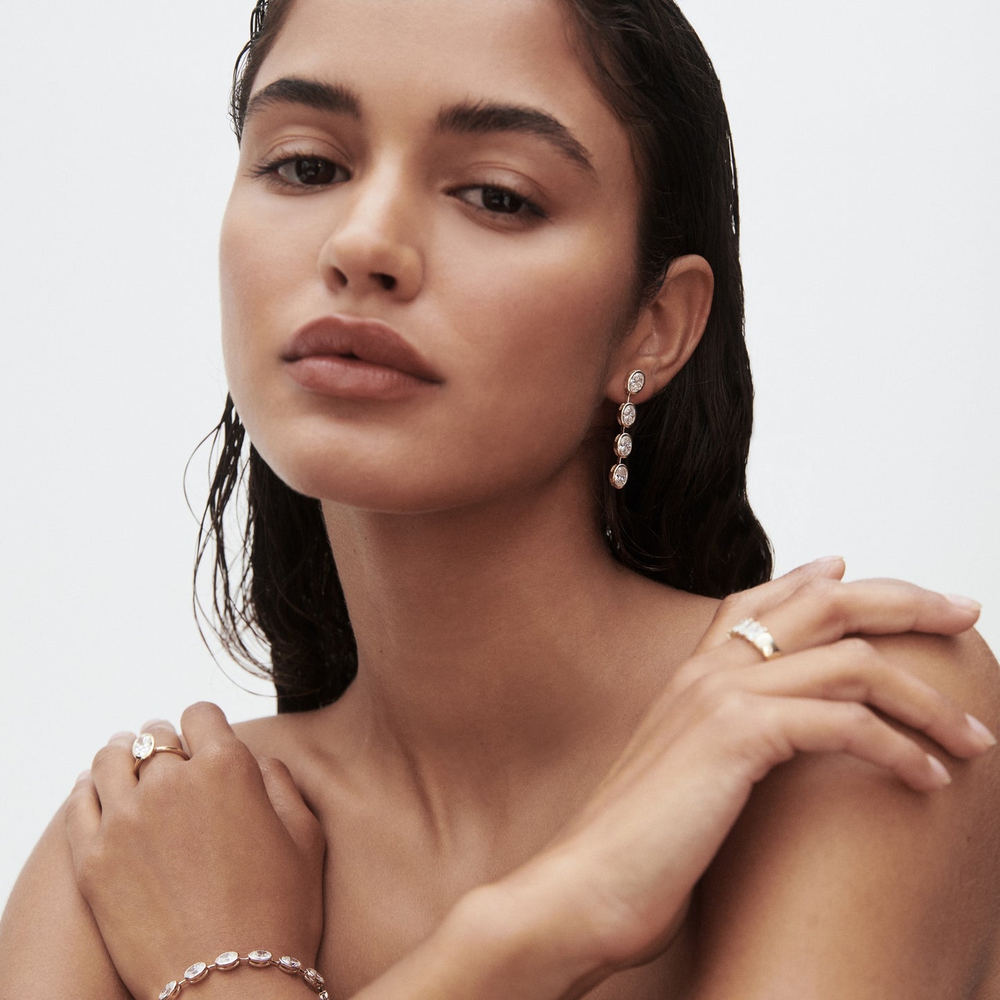 Woman with wet hair wearing luxury jewellery: drop earrings, a bracelet, and rings. Soft lighting, minimalistic background.