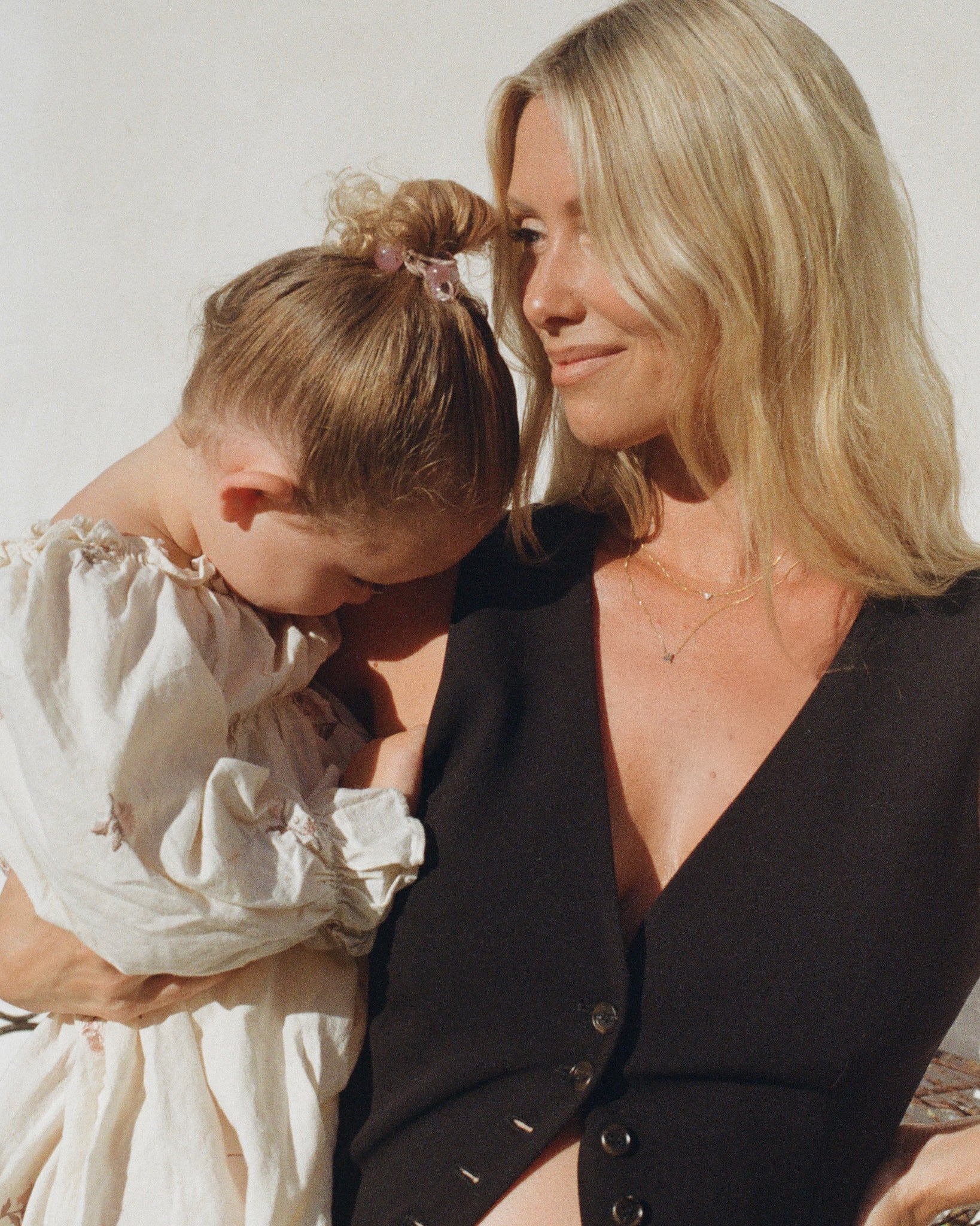 A woman with long blonde hair wears a black top and delicate gold necklace, holding a young child in a ruffled white dress.