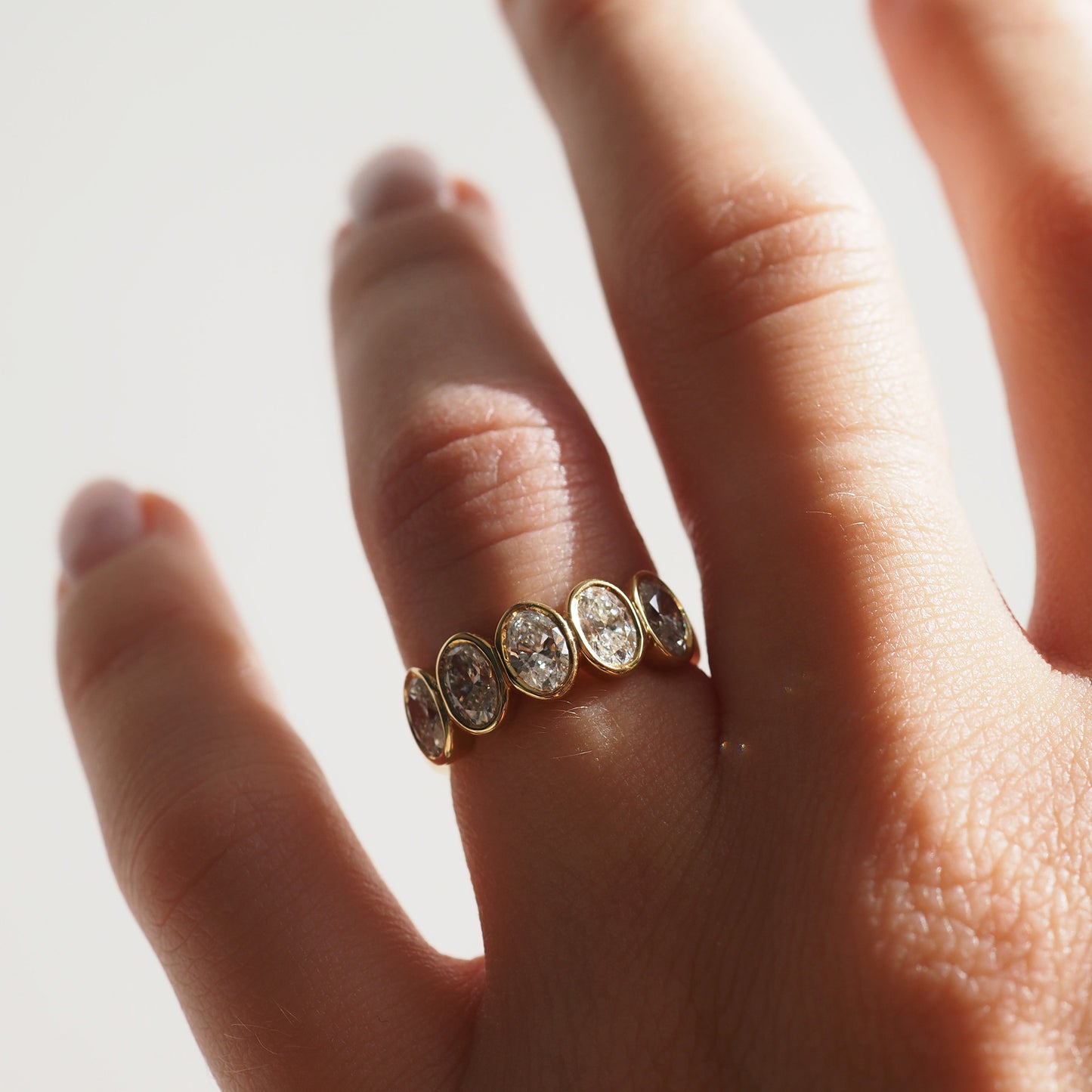 Hand wearing a gold ring with five oval diamonds, each set in individual bezels, against a softly lit background.